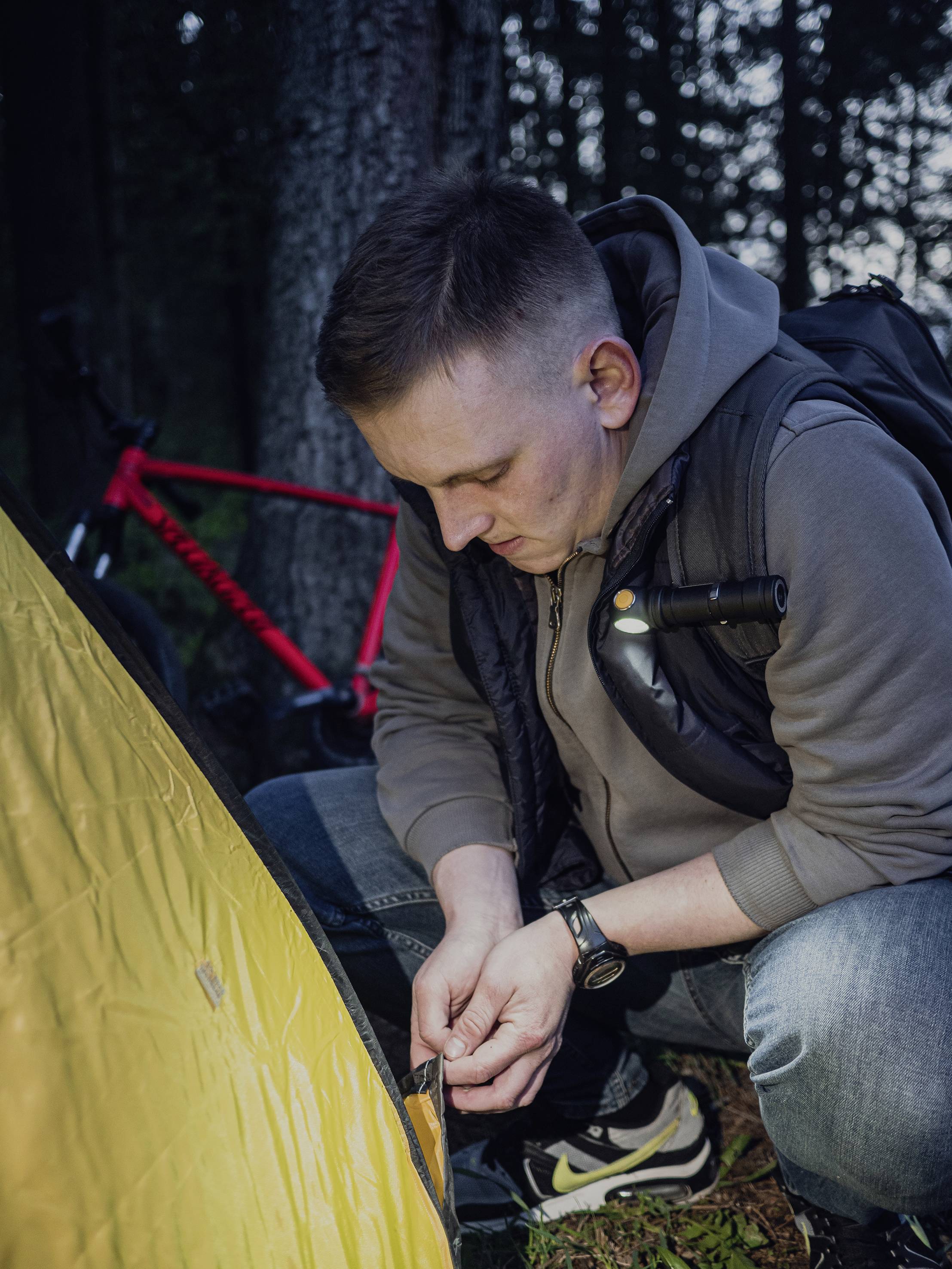 A man is pitching a tent in the woods. A red bicycle is leaning against a tree in the background. It is gradually getting dark.