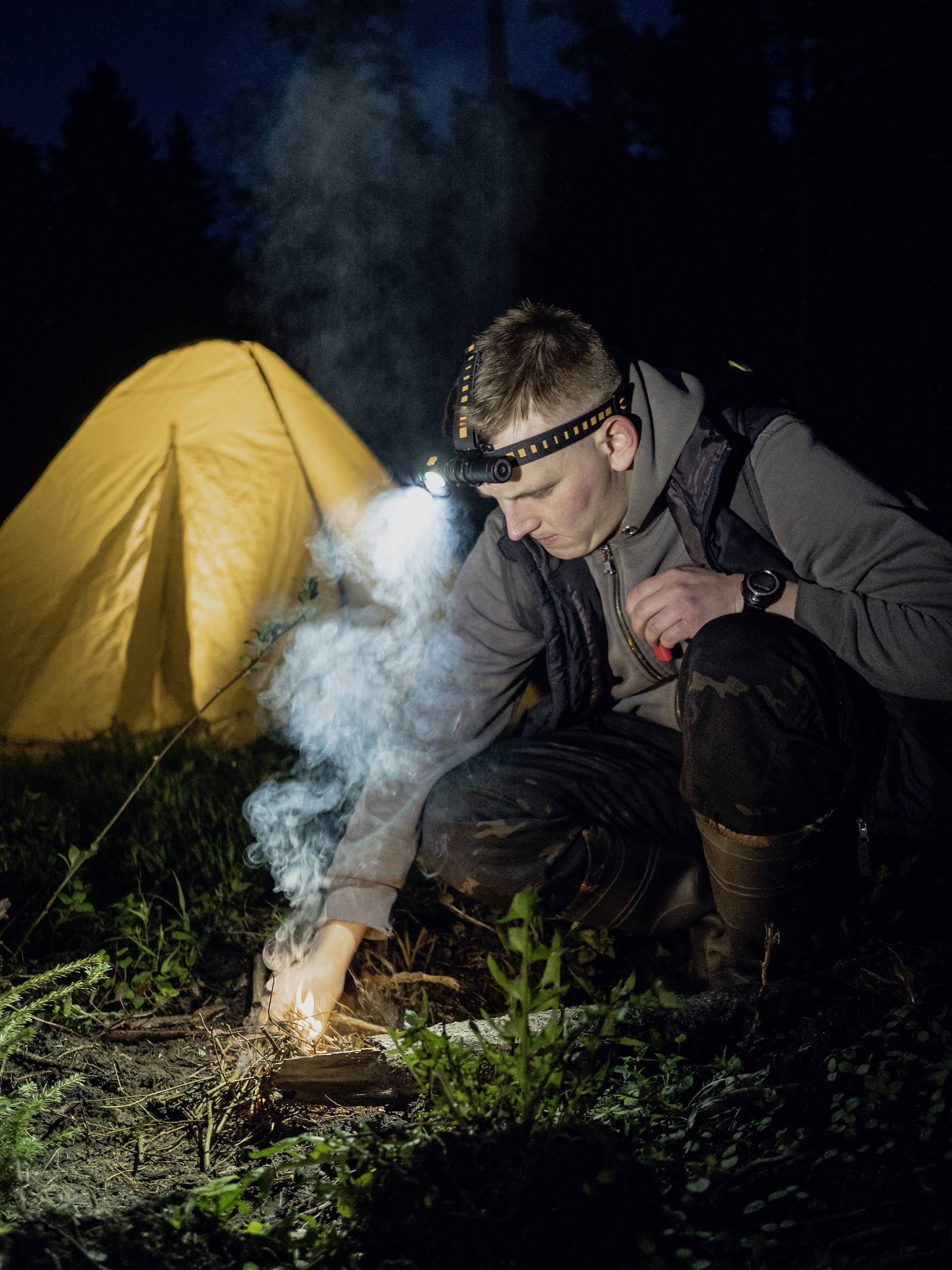 A person wearing a head torch lights a fire in the evening, with an illuminated yellow tent in the background.