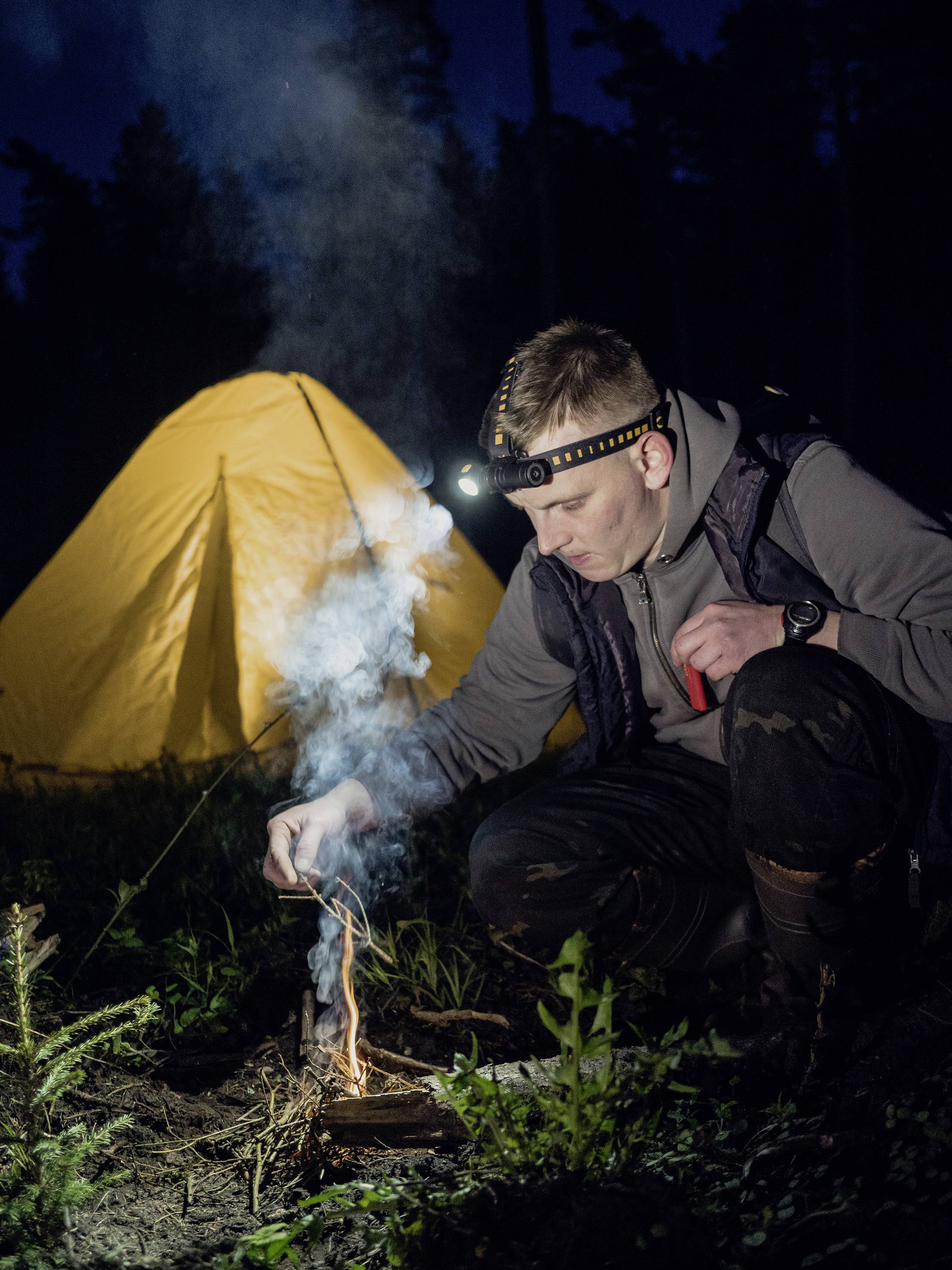 A man wearing a head torch lights a small campfire. In the background, an illuminated yellow tent stands at night in the forest.