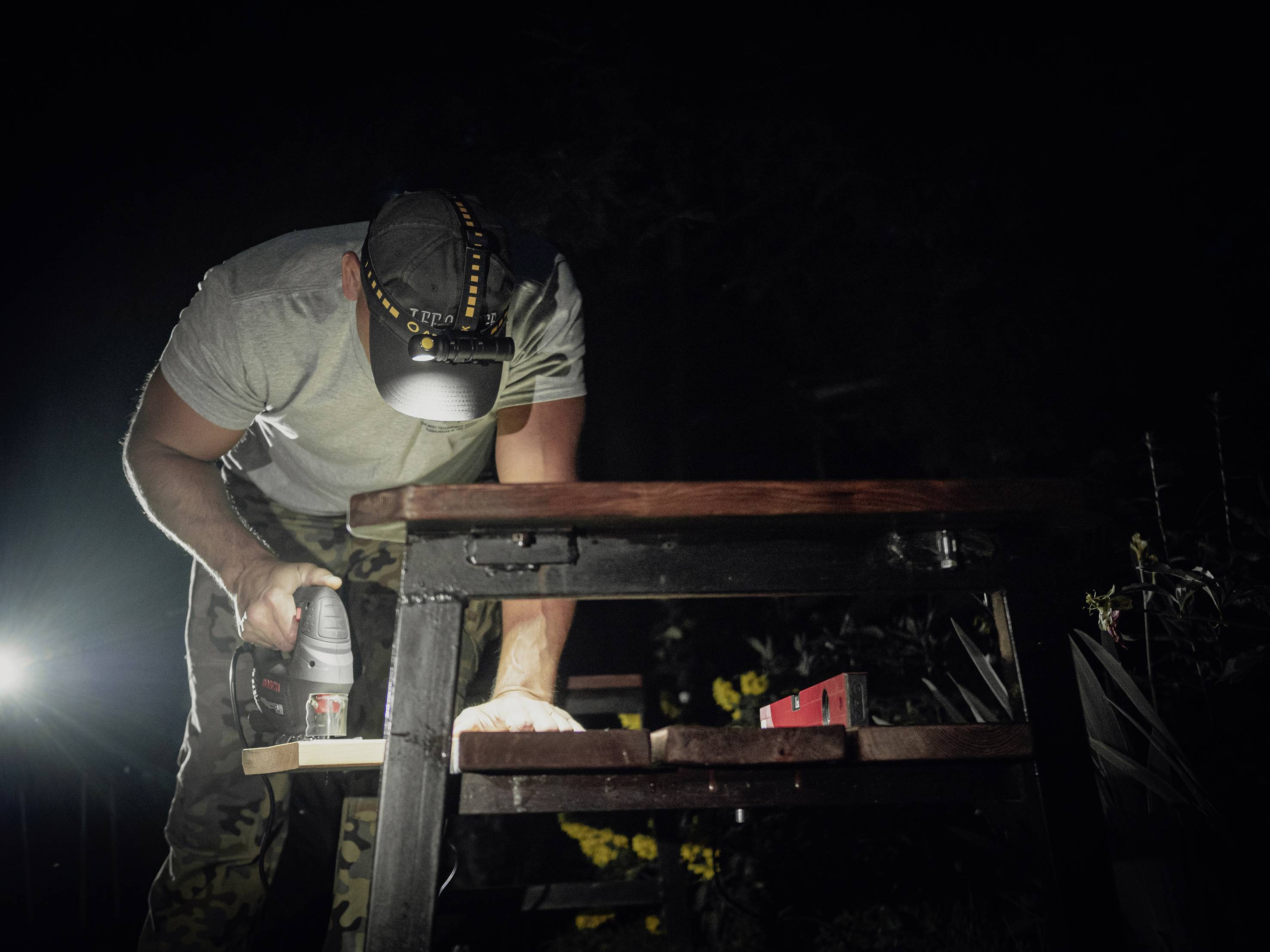 A man is wearing a cap with a head torch, using a drill at a table outdoors, surrounded by darkness.