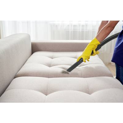 A person is cleaning a beige sofa with a vacuum cleaner. They are wearing yellow cleaning gloves.