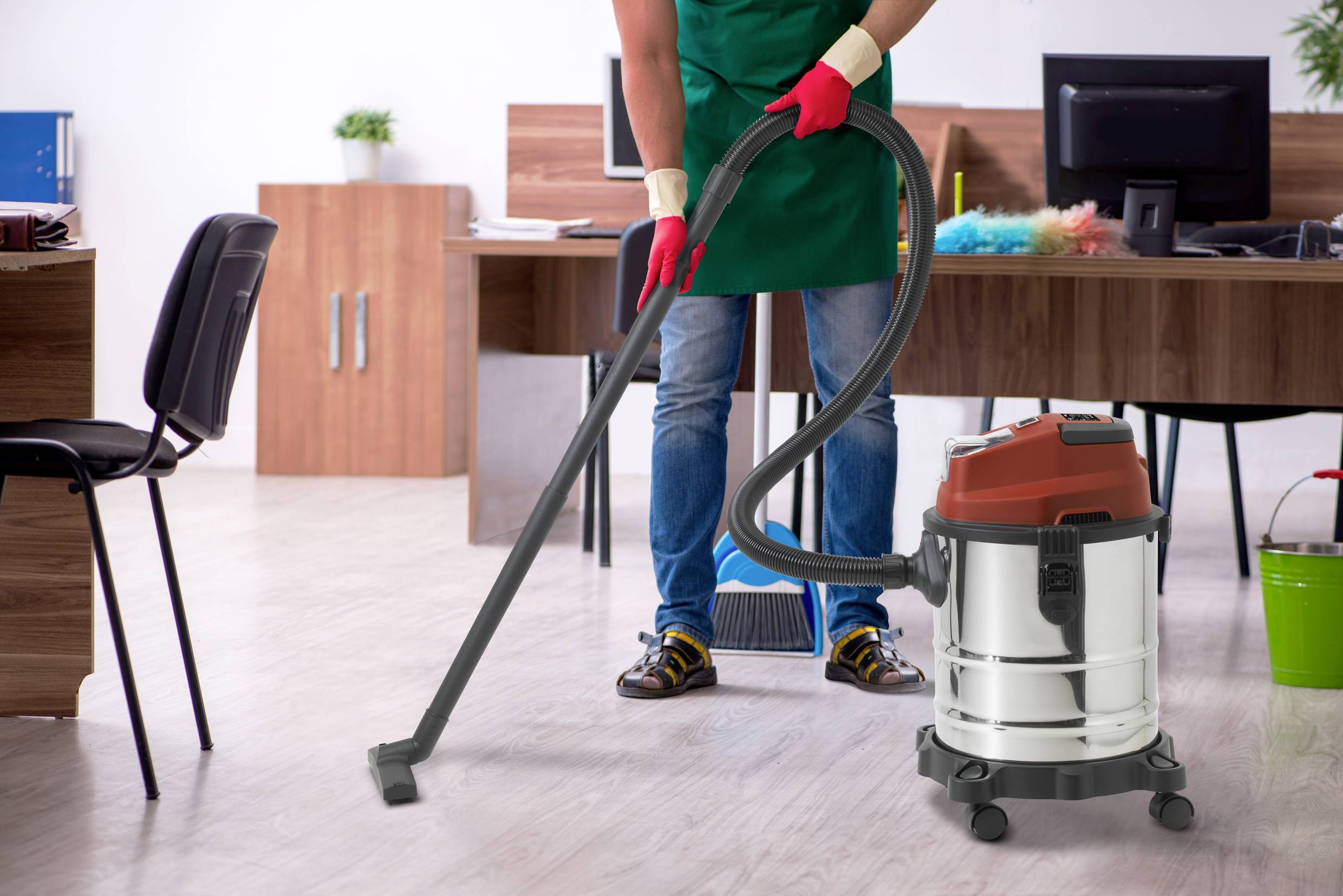 A person is vacuuming an office, wearing red gloves and jeans. In the background, there are wooden desks, chairs, and plants.