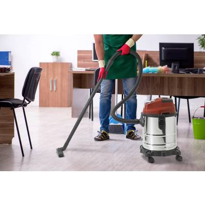 A person is vacuuming an office, wearing red gloves and jeans. In the background, there are wooden desks, chairs, and plants.