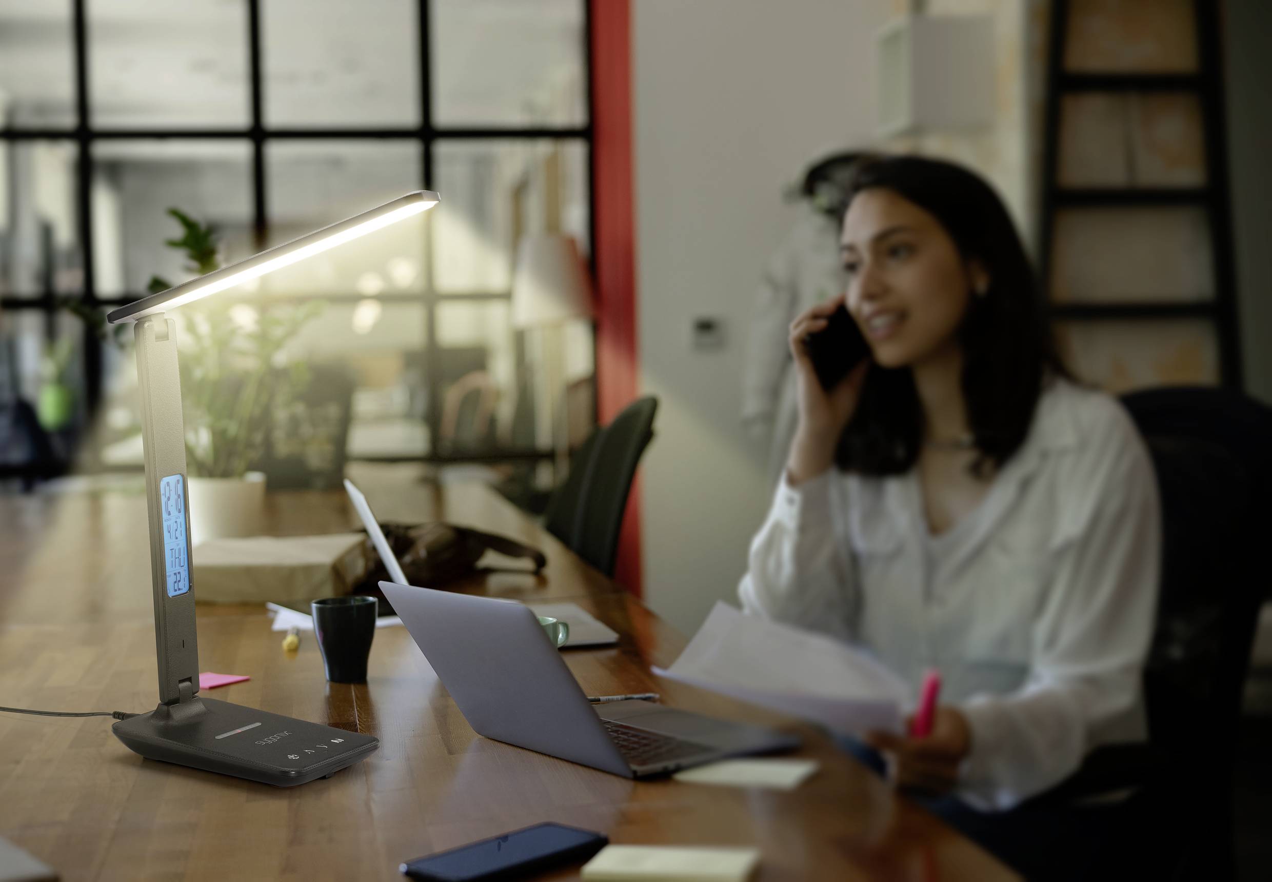 A woman is talking on the phone at a desk with a laptop and documents, a desk lamp illuminating the space. She appears busy and focused.