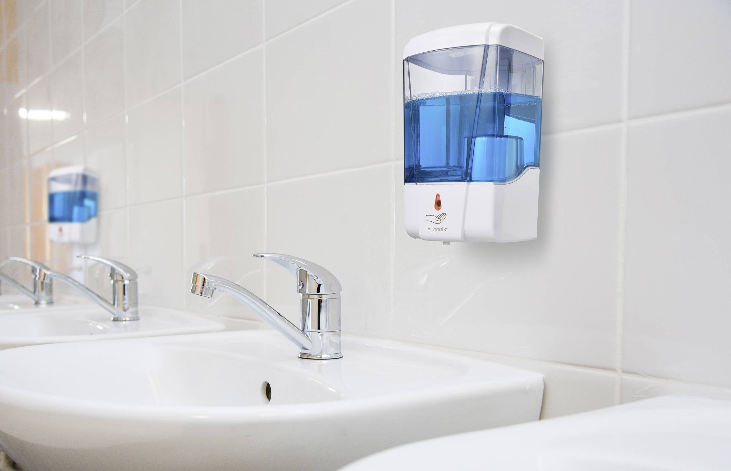 A washbasin area with multiple white ceramic sinks and wall-mounted soap dispensers with blue liquid on white-tiled walls.