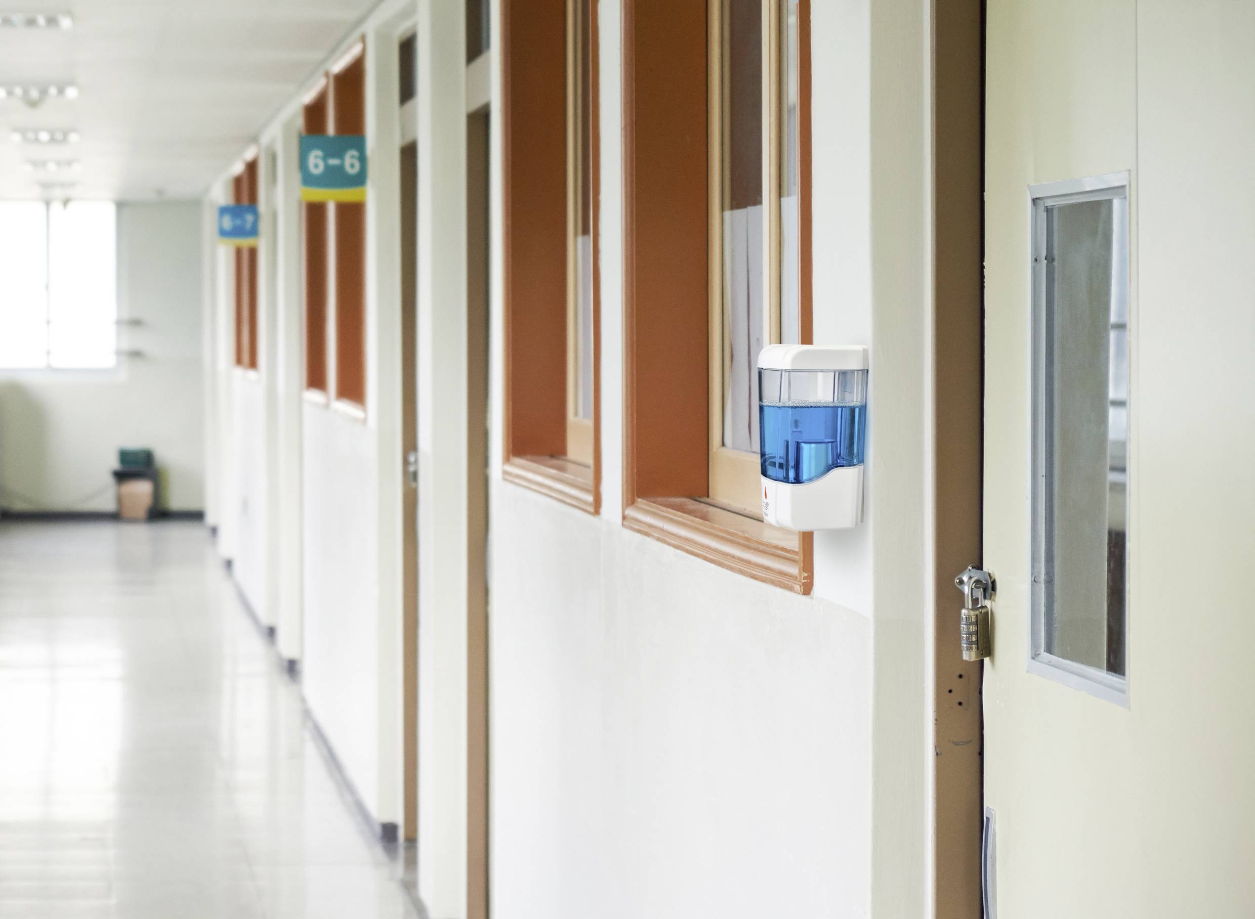 Long corridor with several closed doors on the right-hand side, numbered signs (6–6) above each room, hand sanitiser dispensers on the wall.