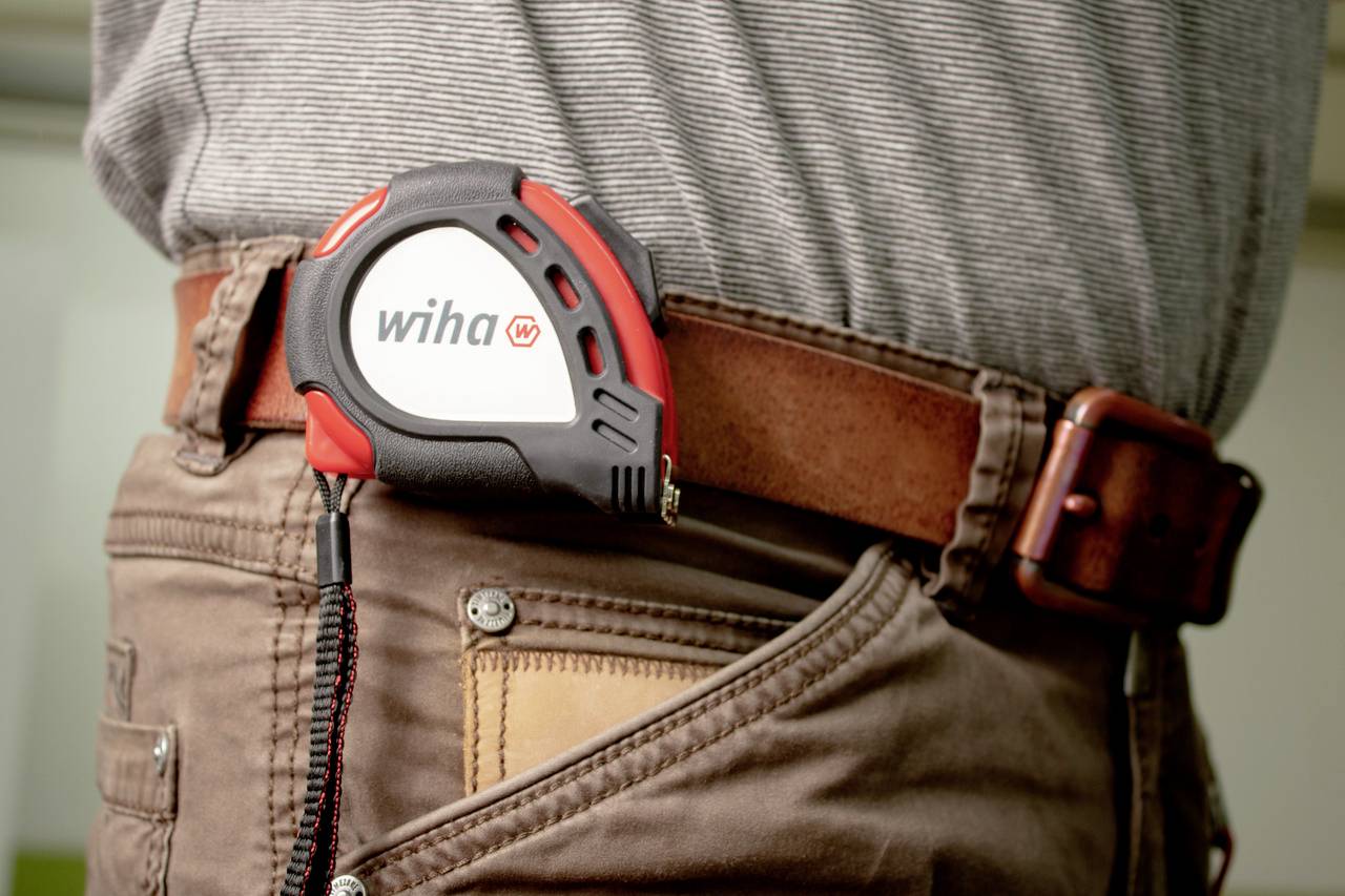 A person is wearing a tape measure on their belt. The tape measure has a grey and red casing with the 'Wiha' logo.
