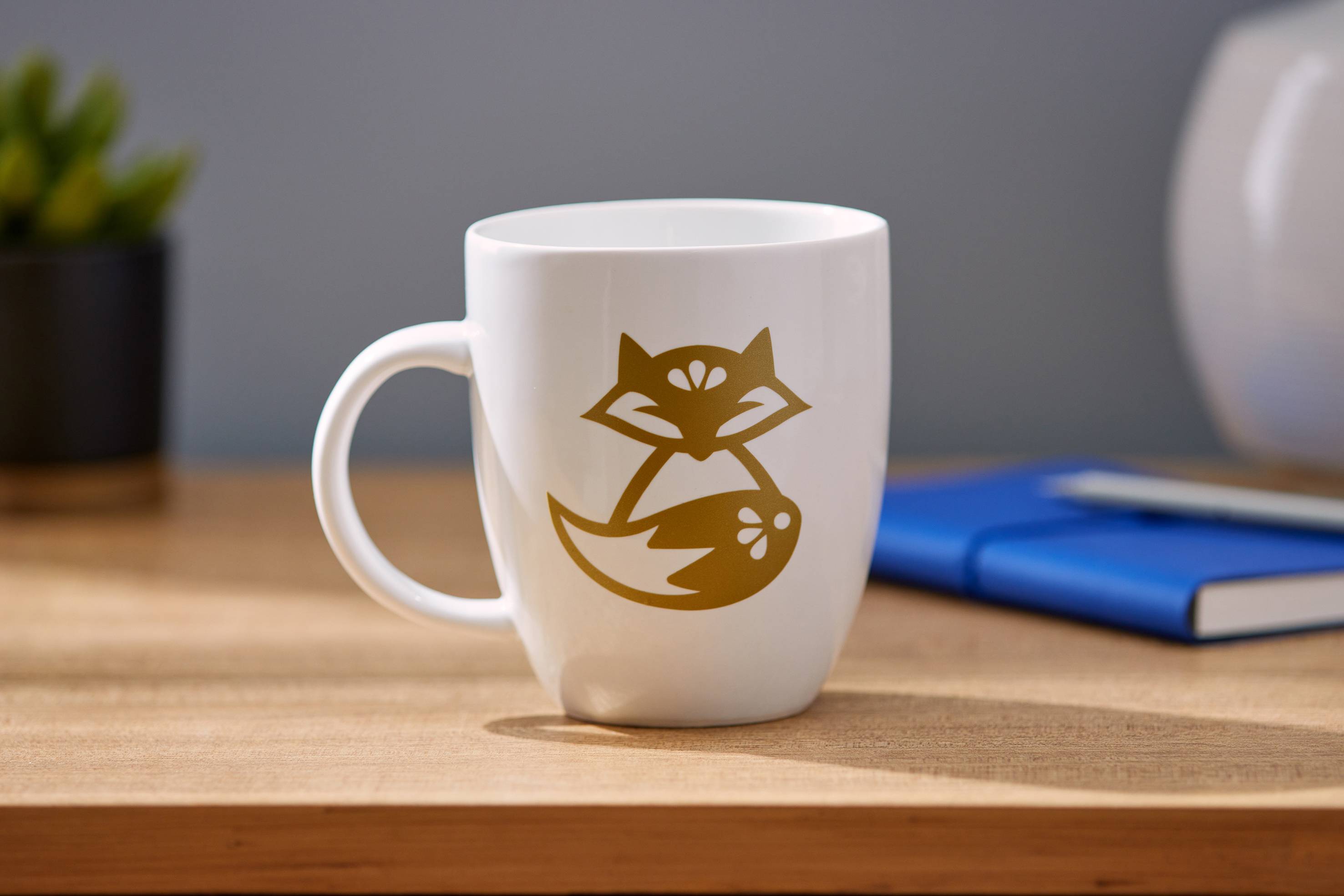A white coffee cup with a golden fox design sits on a wooden table. In the background, a notebook and a plant can be seen.