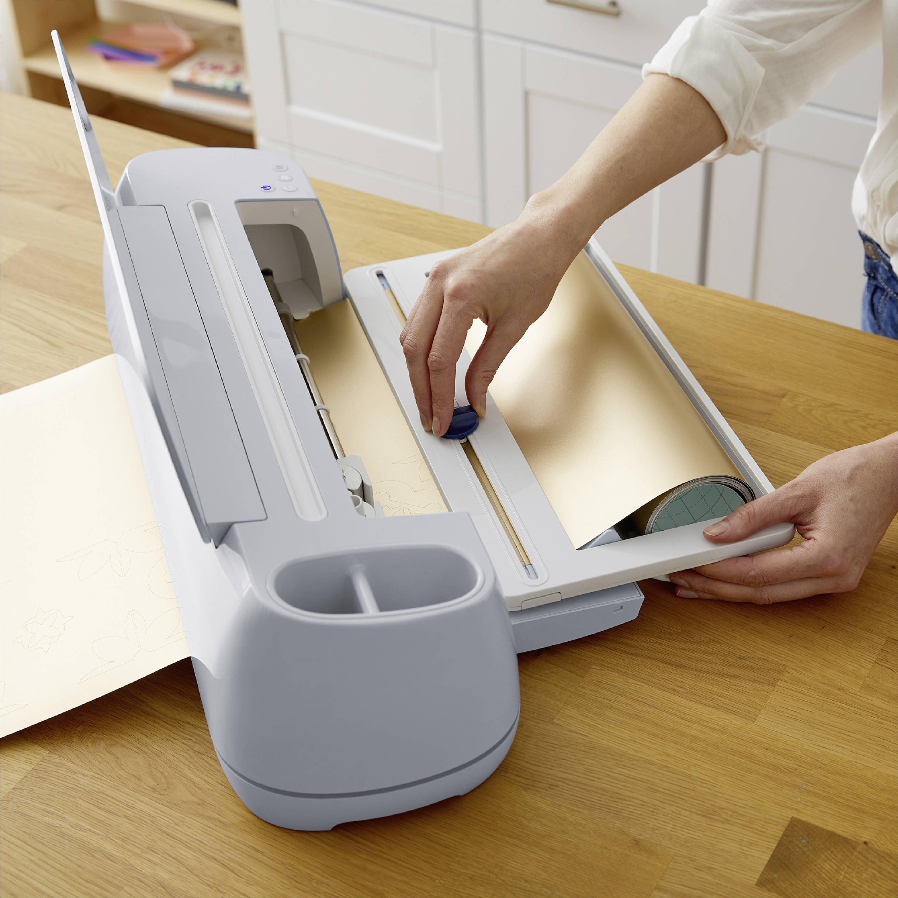 A person is operating a cutting plotter on a wooden table. The plotter is cutting a design out of a gold foil sheet.