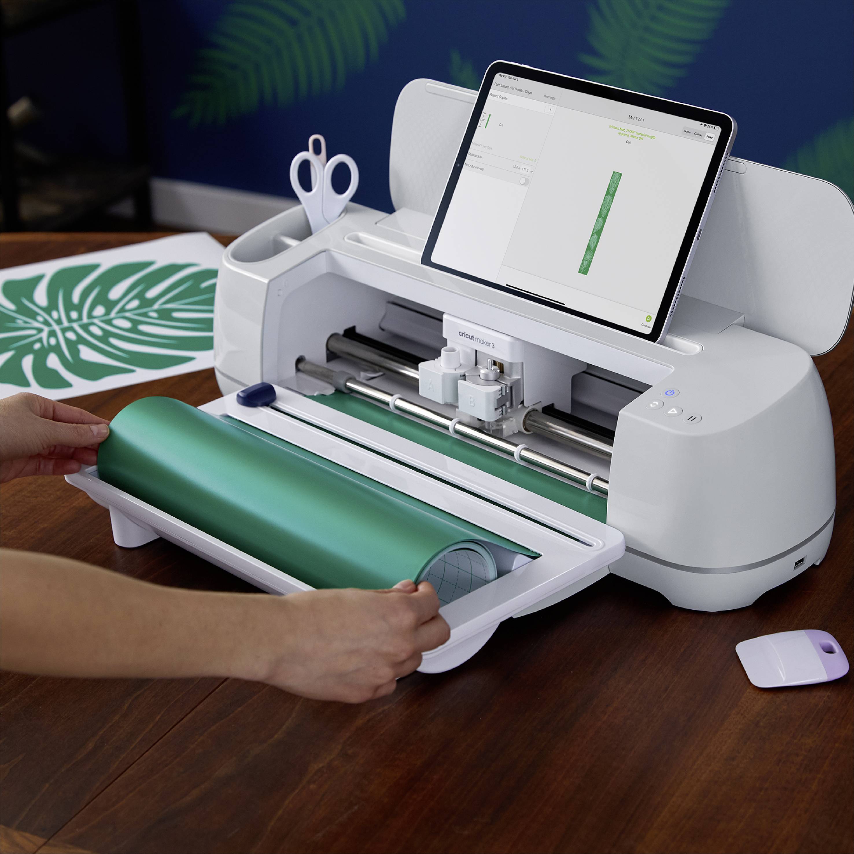 A cutting plotter sits on a table cutting green vinyl. A tablet displays a leaf design. A hand is holding the vinyl roll.