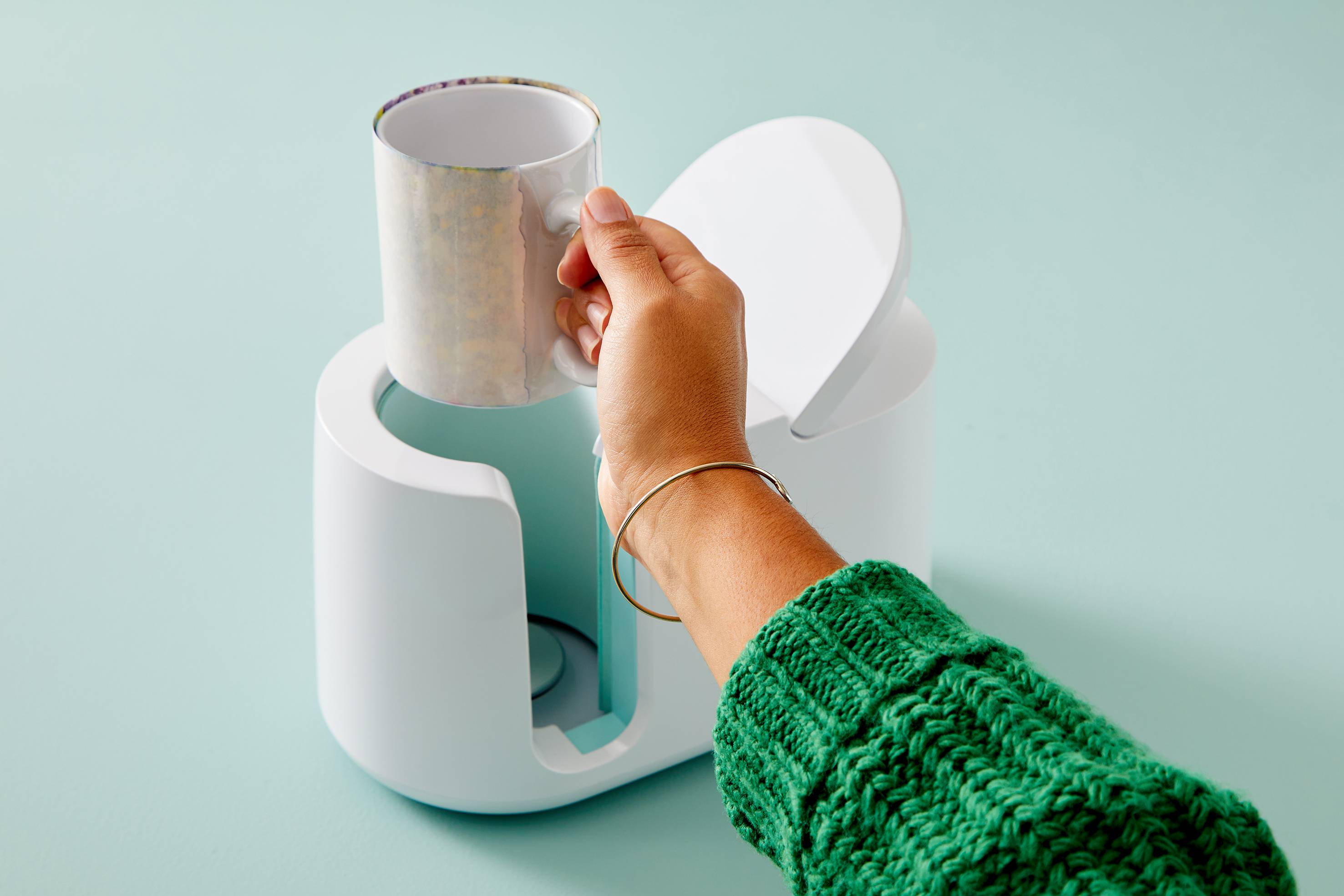 A hand in a green jumper places a cup into a white, open coffee machine against a mint green background.
