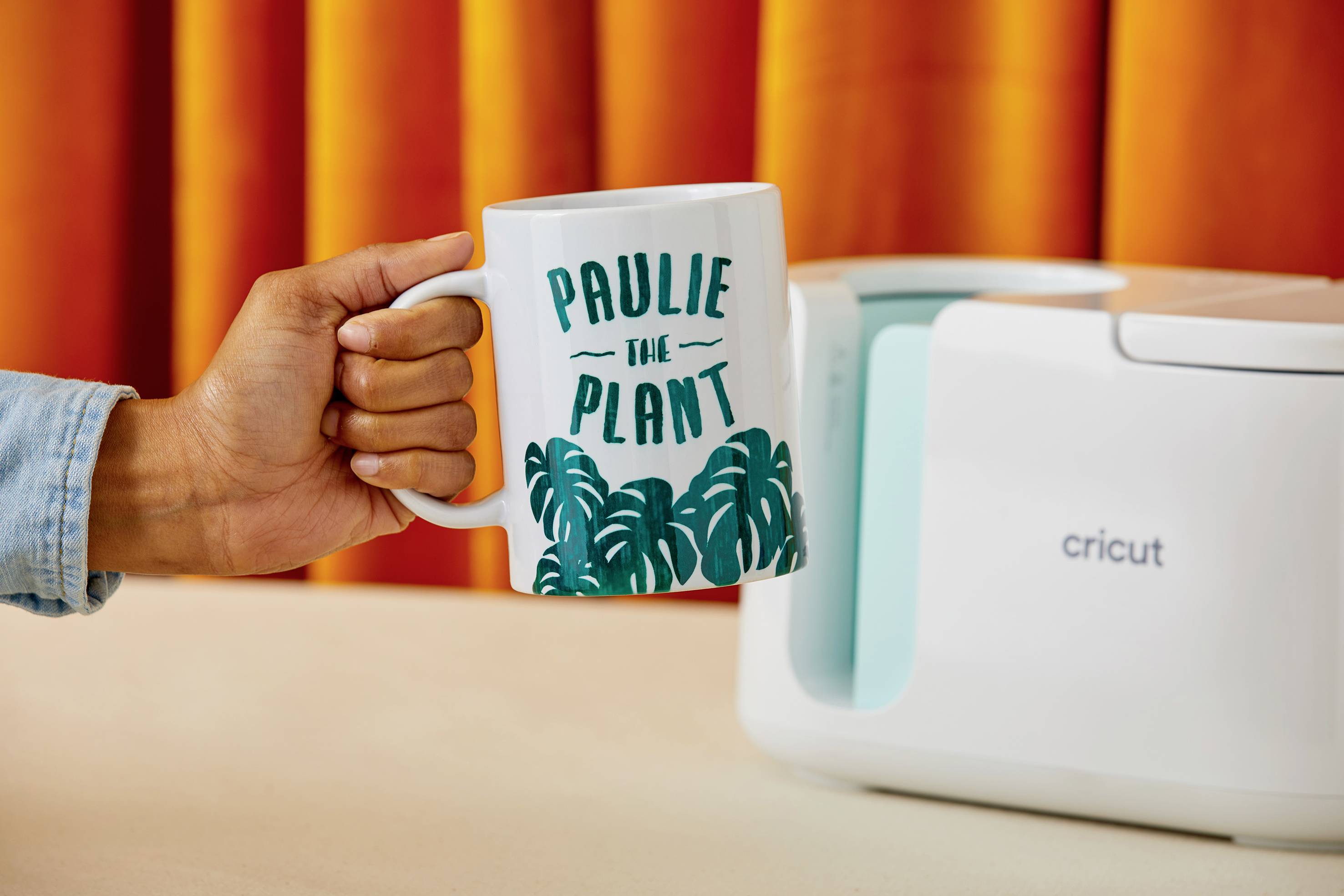 A hand holds a mug printed with 'Paulie the Plant' in front of a white device on a table, with an orange curtain in the background.