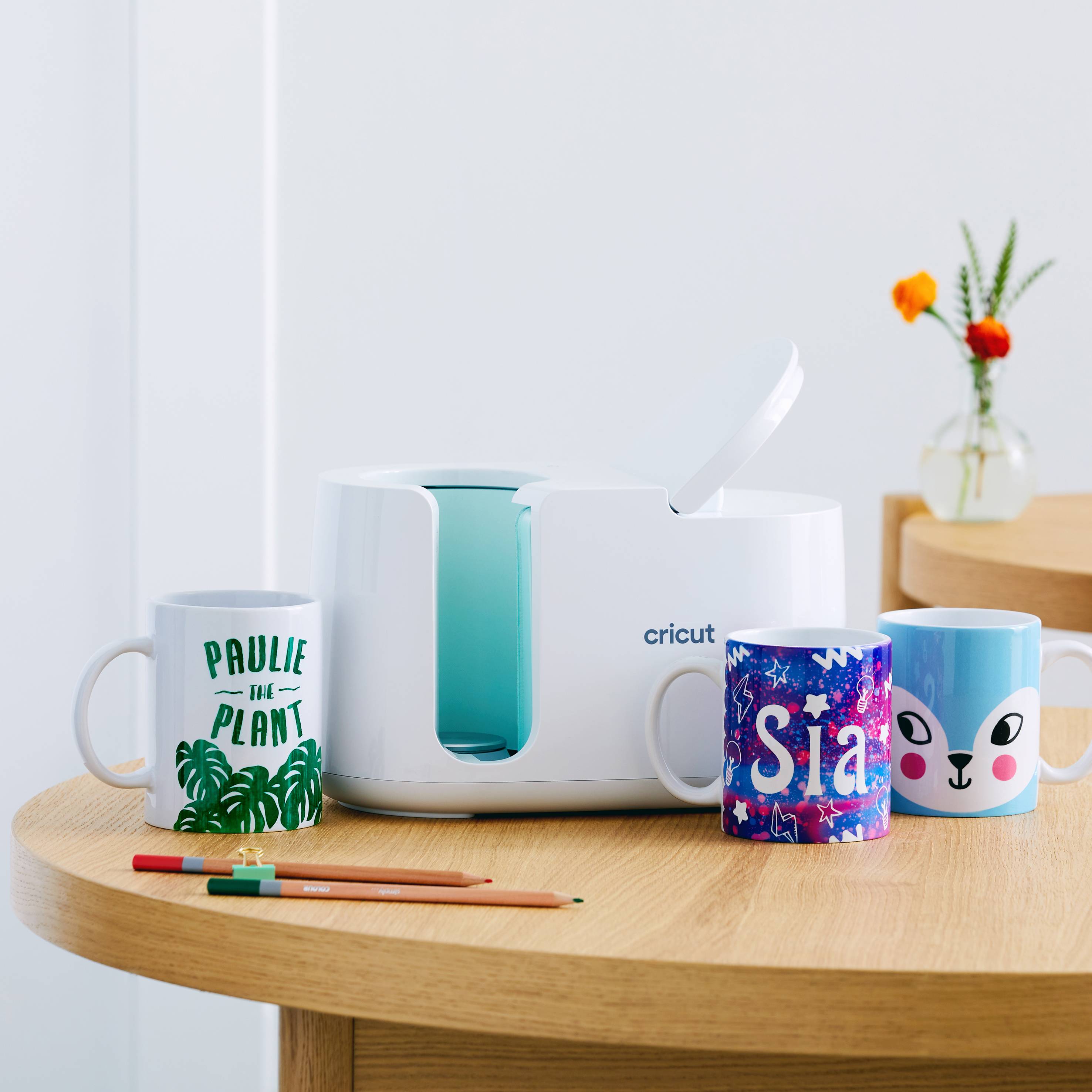 A device with a 'Cricut' logo on a table, surrounded by three mugs with different designs and two pens in the foreground.