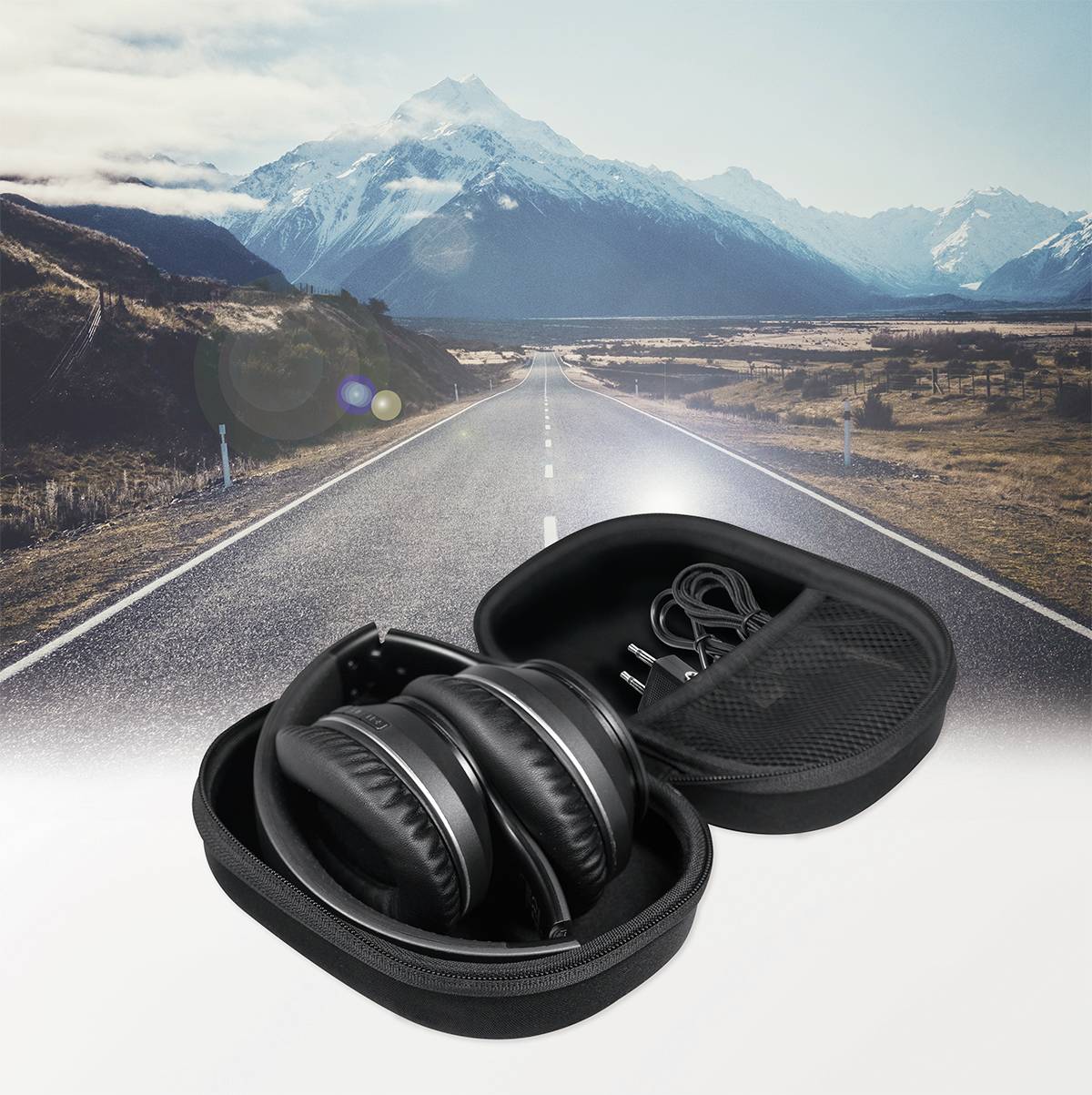 A pair of black headphones lies in an open case on a road with snow-capped mountains in the background.