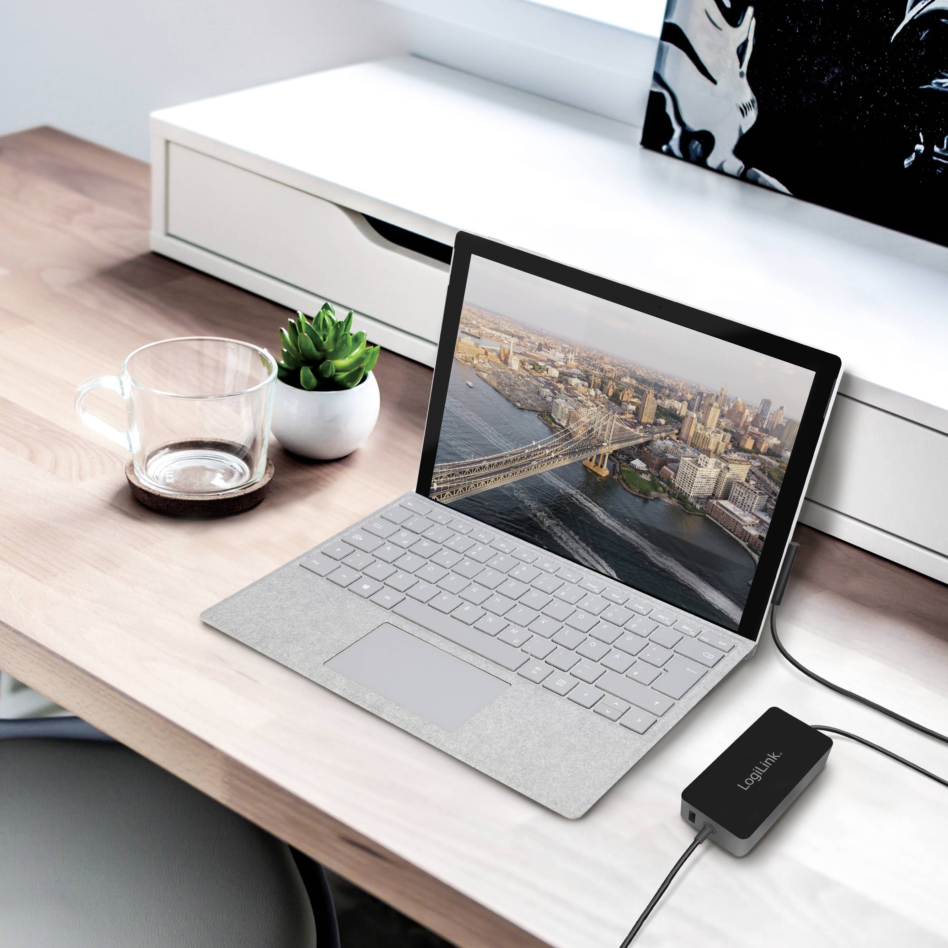 A laptop on a desk displays an image of a cityscape. Next to it are an empty glass mug and an electronic device.
