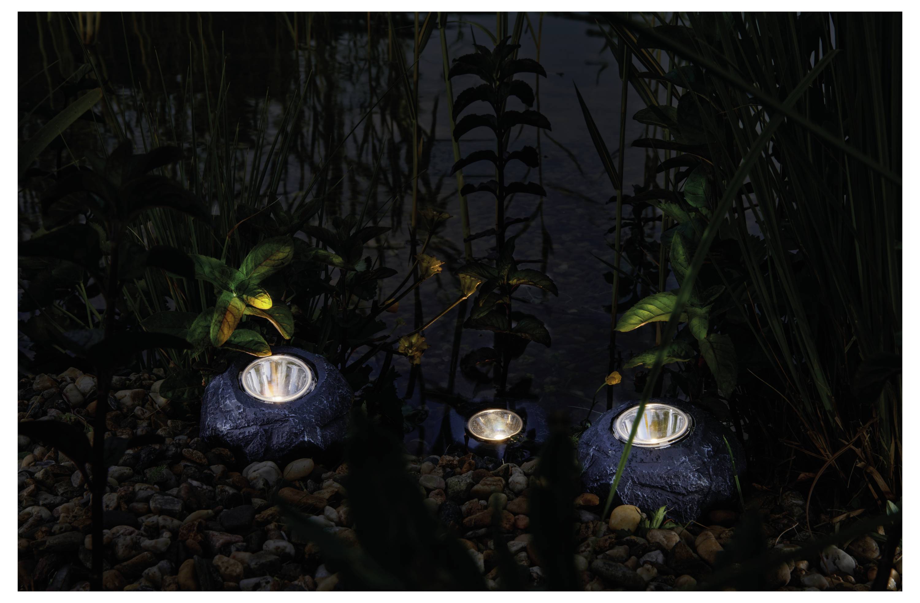 Three solar lights resembling rocks illuminate a garden path surrounded by plants and pebbles at night, creating a serene atmosphere.