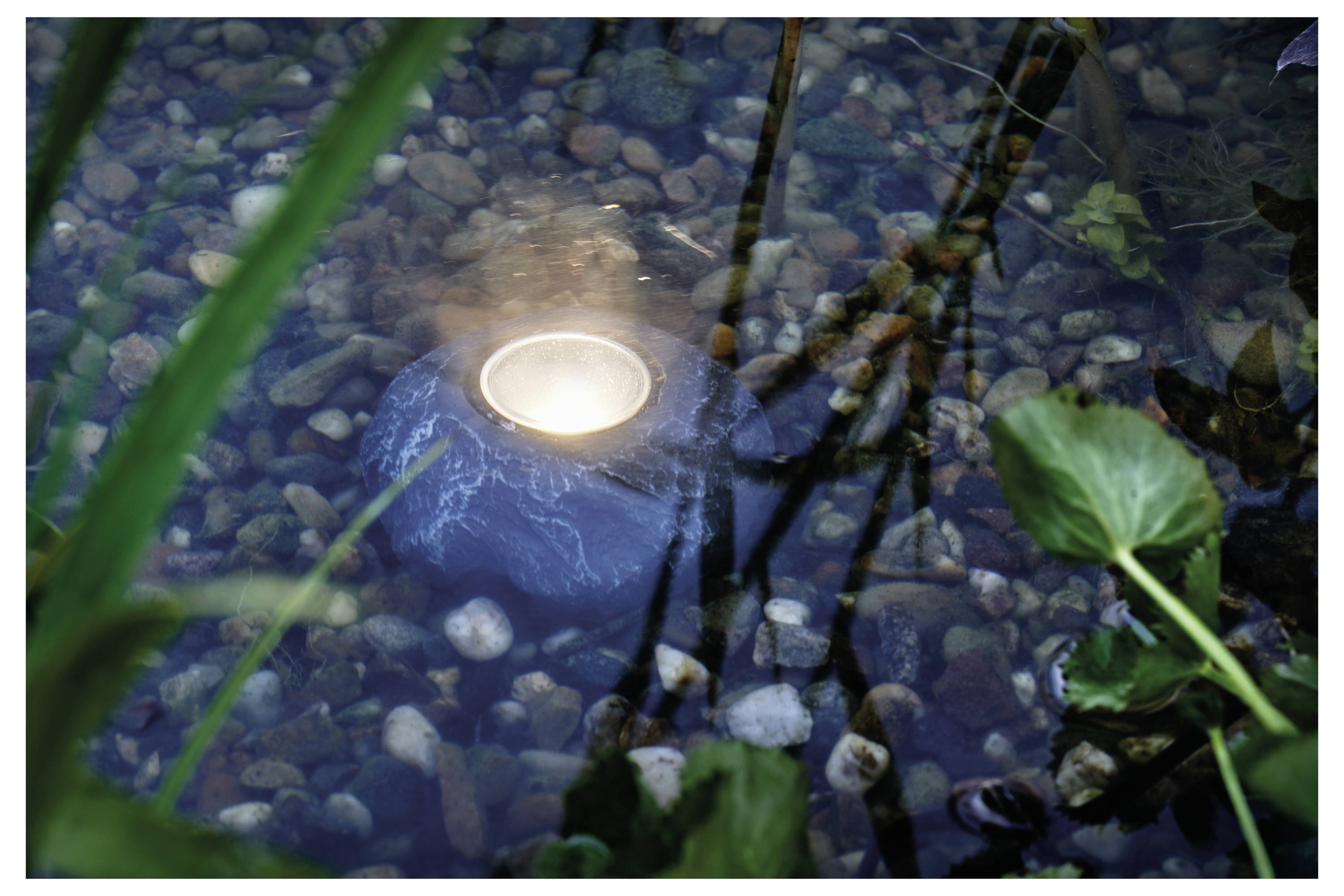 A candle in a stone holder floats in a shallow, rocky pond with greenery around, creating a serene and calming atmosphere.