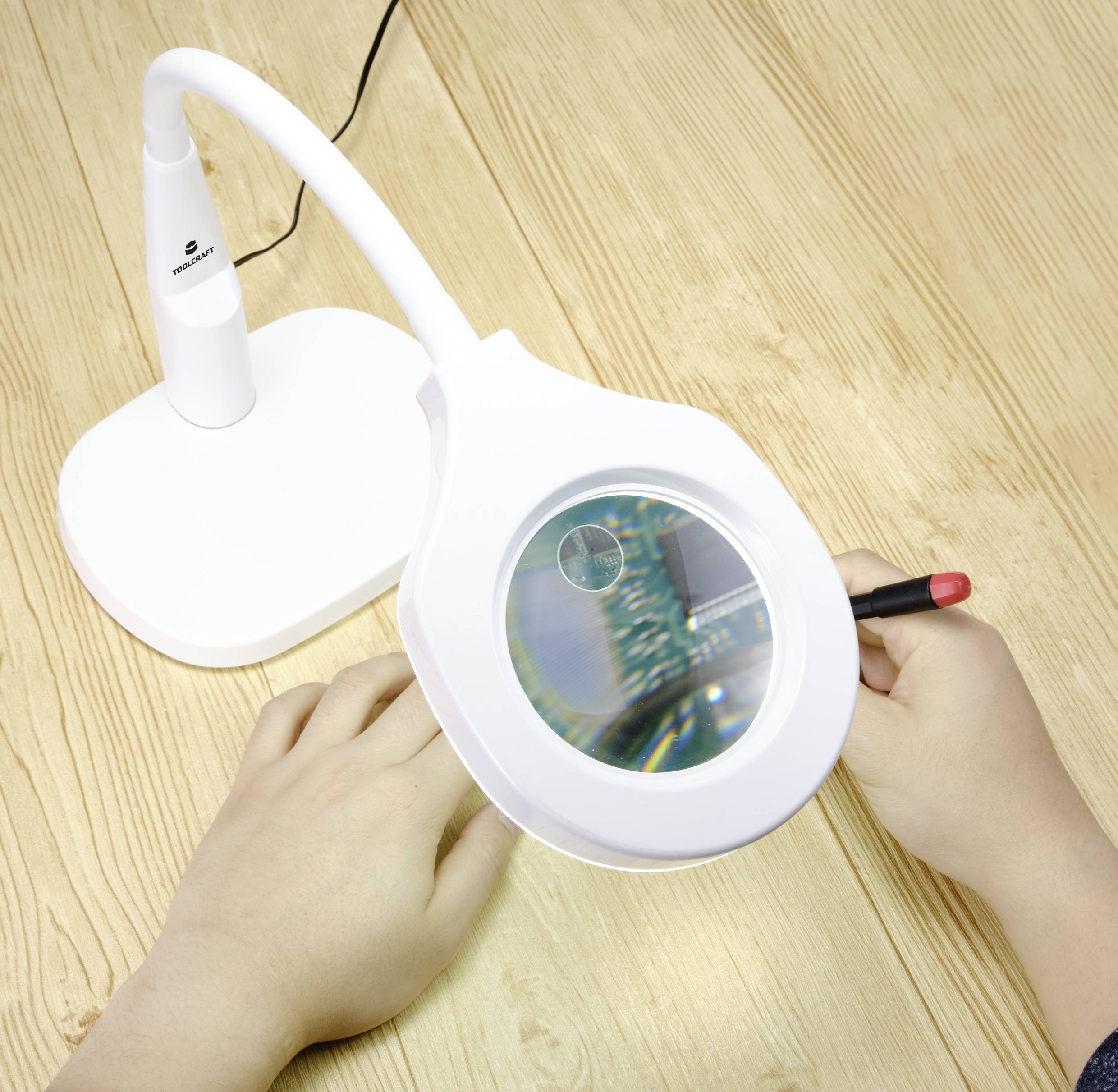 Two hands are holding a red soldering station, using a magnifying glass to closely examine a circuit board on a wooden table.