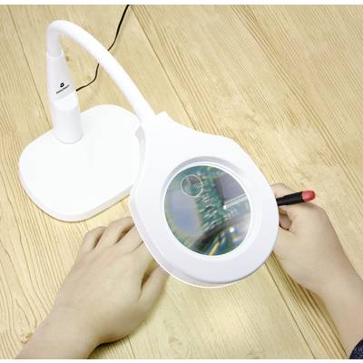 Two hands are holding a red soldering station, using a magnifying glass to closely examine a circuit board on a wooden table.