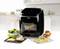 A black air fryer sits on a kitchen worktop, surrounded by a salad bowl, two ready-made pizzas and muffins.