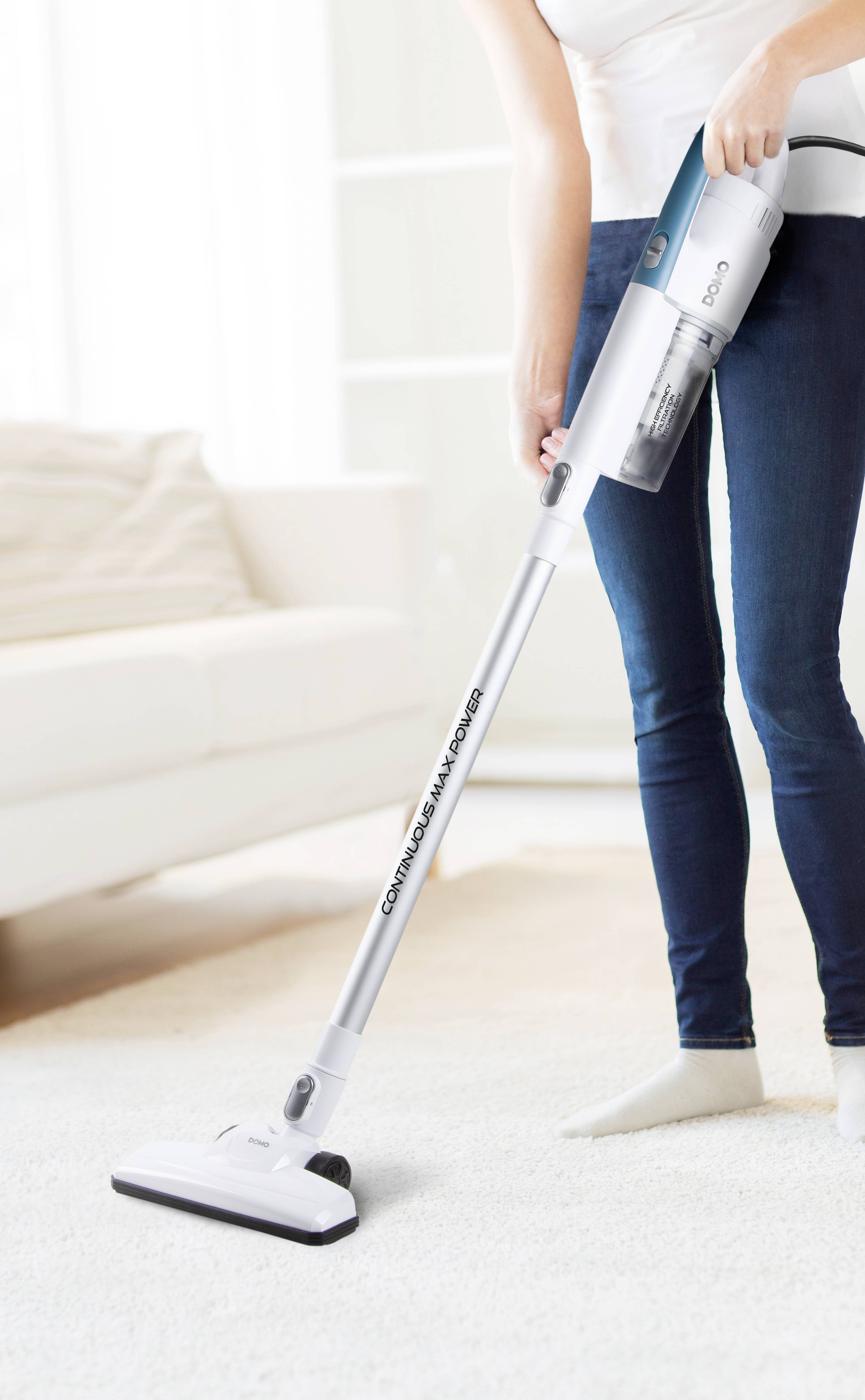 A person is vacuuming a white carpet in a bright room with a white, cordless vacuum cleaner.