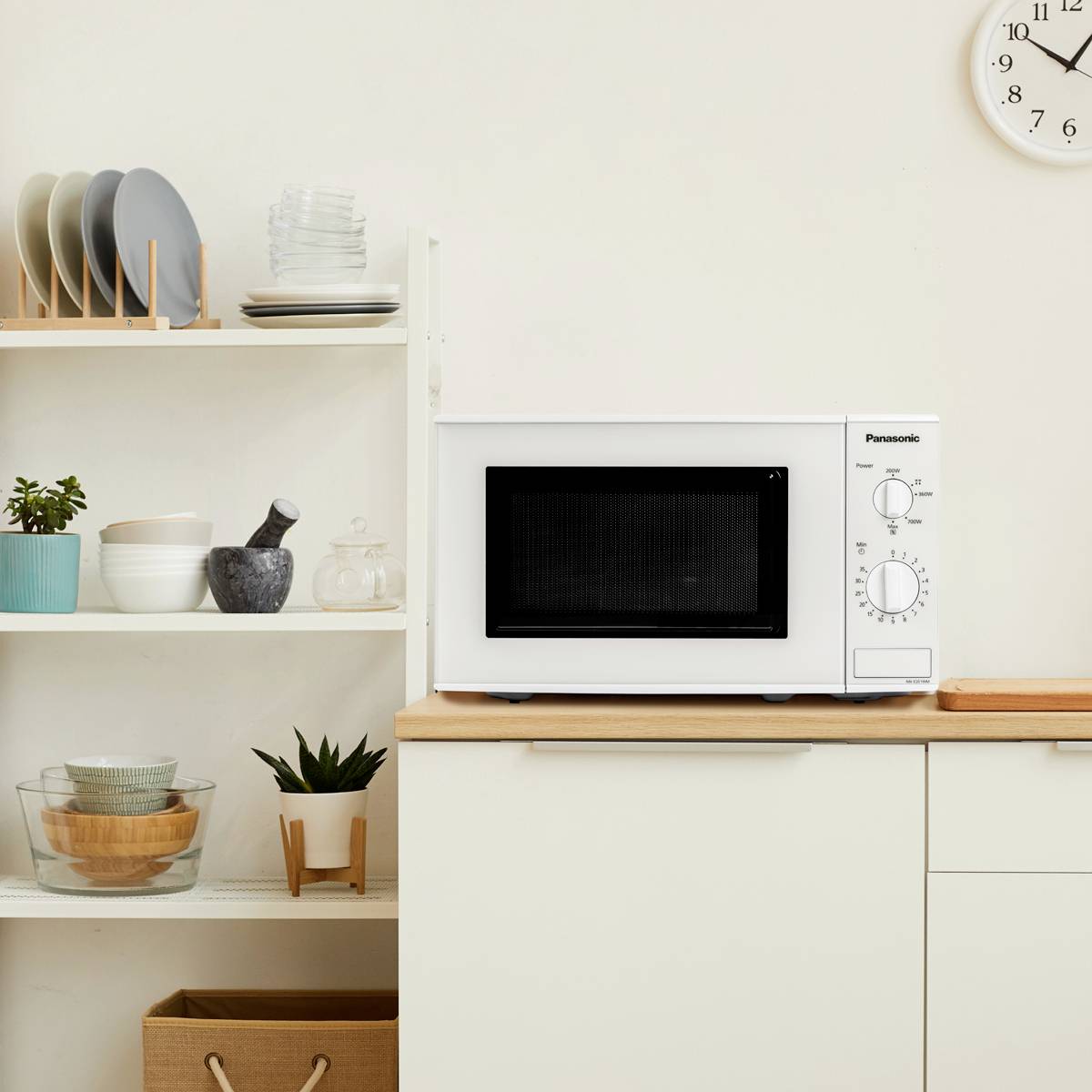A microwave sits on a kitchen worktop, surrounded by shelves of crockery, pots and decorative items. A clock on the wall shows 10:10.