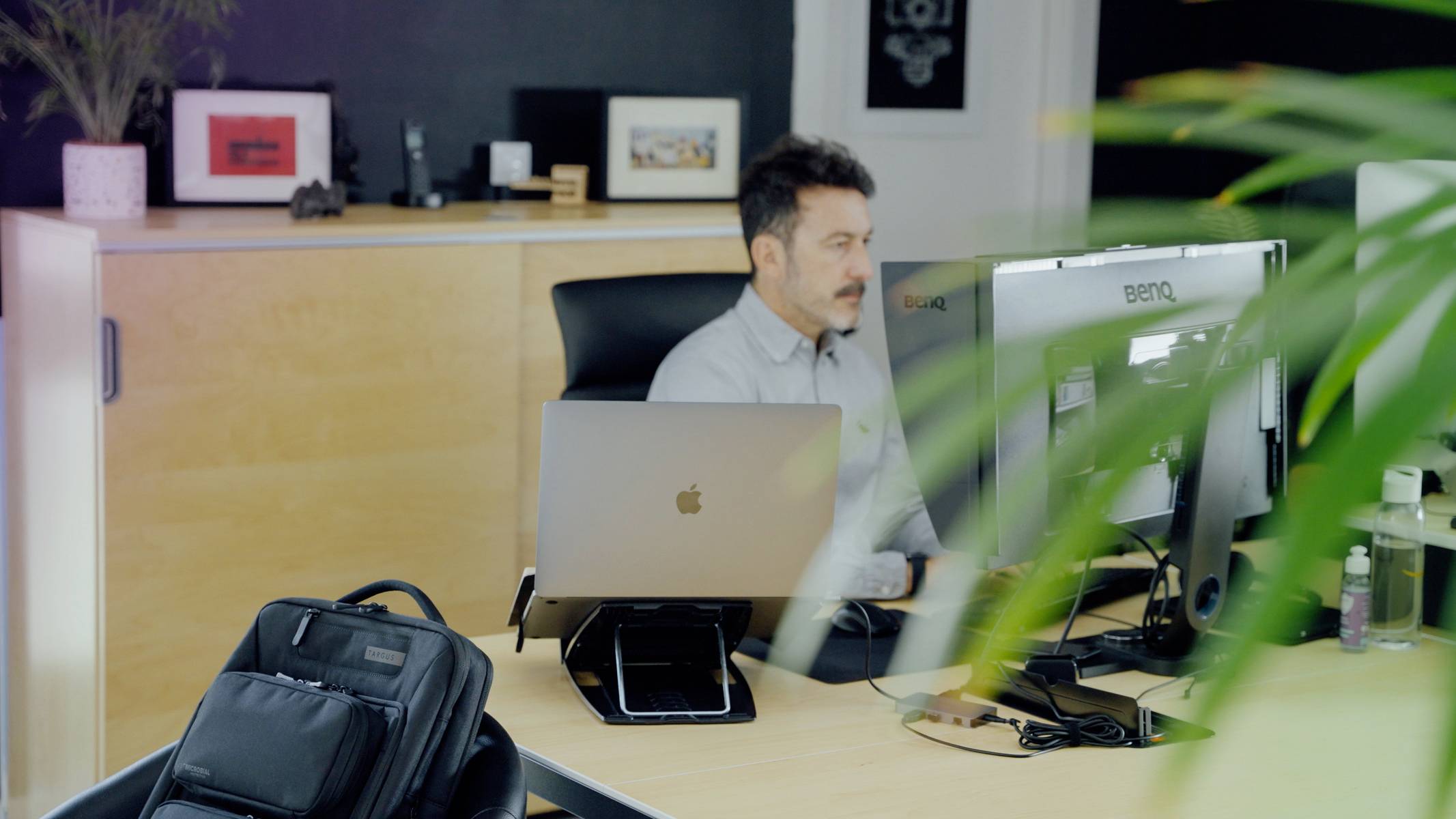 A man sits at a desk with two monitors. He is working intently, with an office in the background, and a plant in the foreground.