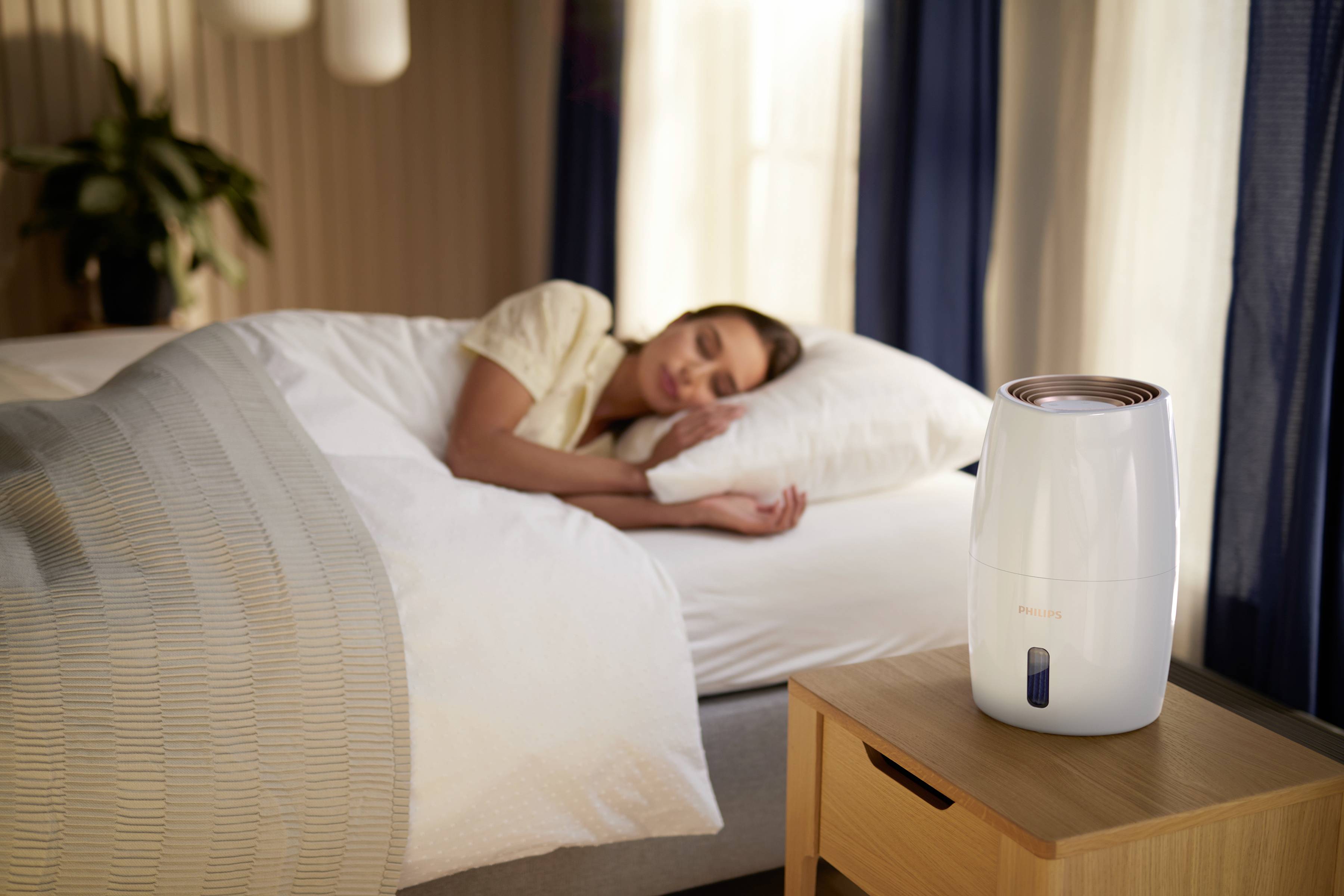 A woman is sleeping in a bed next to a humidifier on the bedside table. The room is cosy and well-lit.