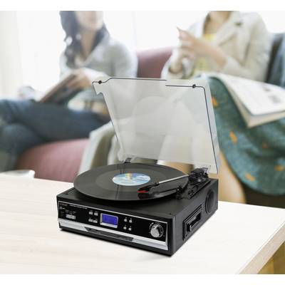 A modern record player with a vinyl record placed on it stands on a table in the foreground, with two people sitting blurred in the background.