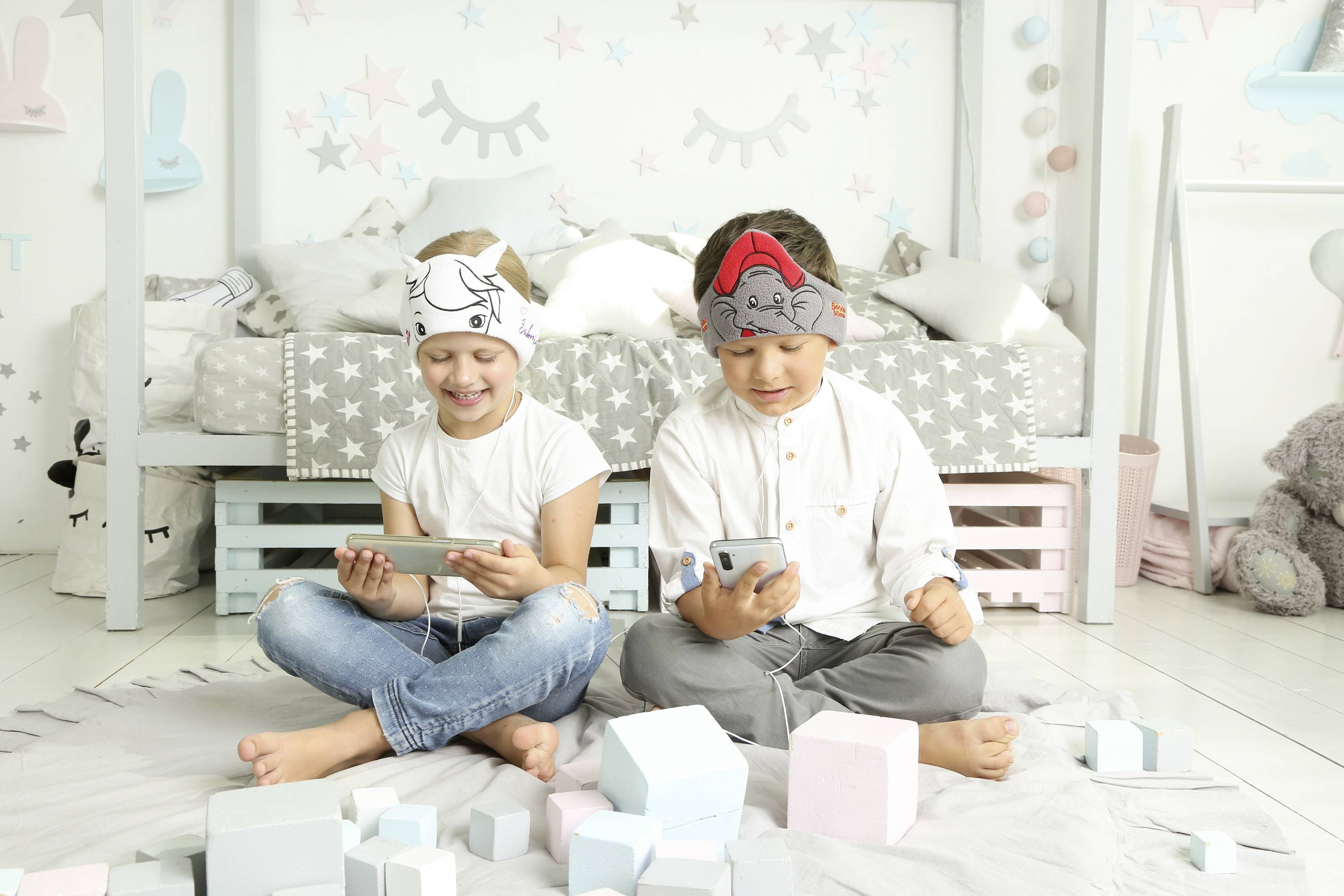 Two children are sitting on the floor and playing with smartphones. They are wearing headwear with animal motifs. Background: a child-decorated room.