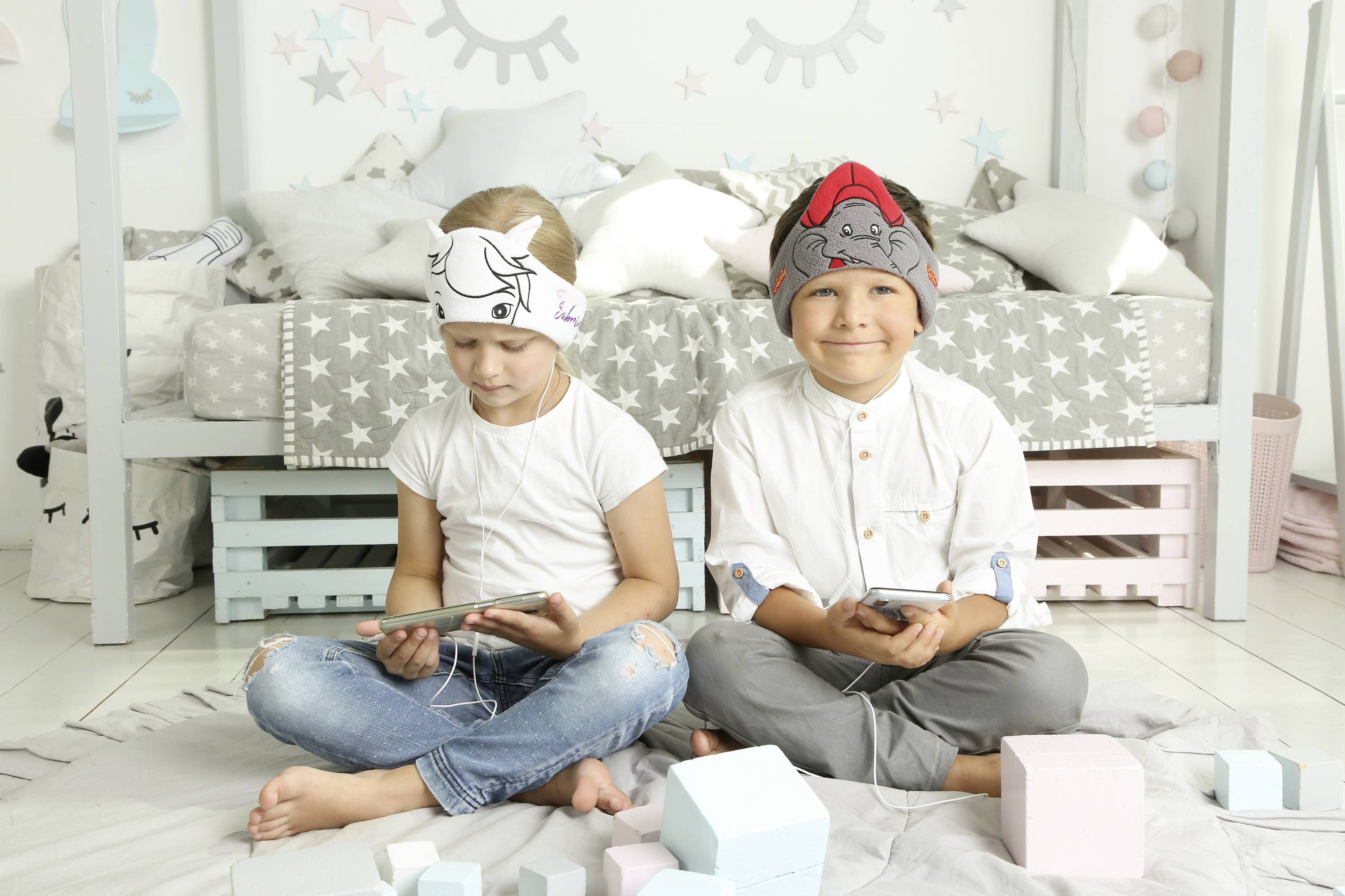 Two children are sitting on the floor in a children's bedroom, both wearing headbands and holding tablets. A bed is visible in the background.