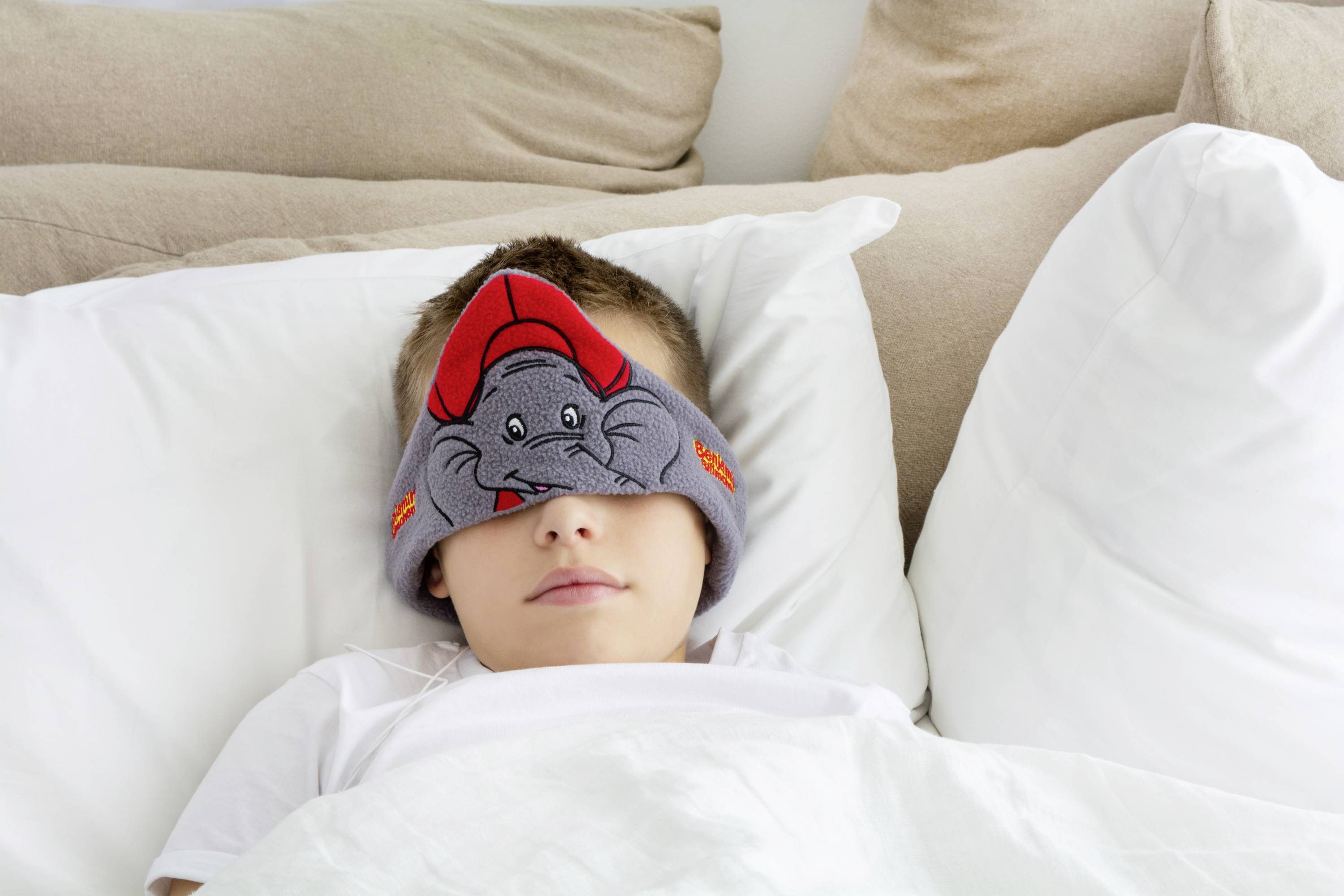 A child is sleeping in bed wearing a sleep mask with an elephant design. They are lying between white pillows and under a white duvet.