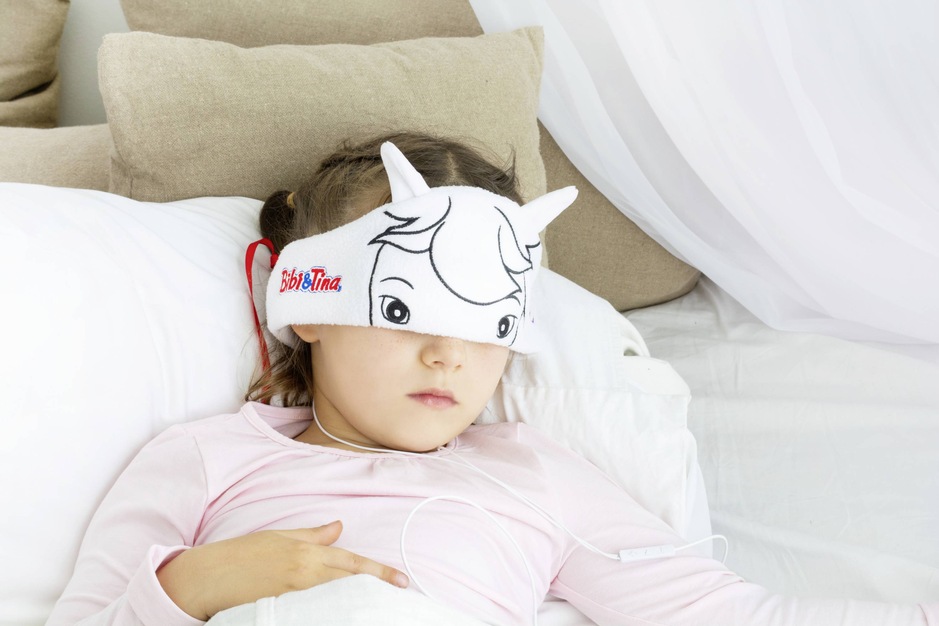 A child is lying in bed wearing a cartoon-themed sleep mask. They appear to be relaxing or sleeping.