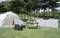 Tent and camping equipment on a green meadow, surrounded by trees. Table with food, solar panel, and cool box in the foreground.