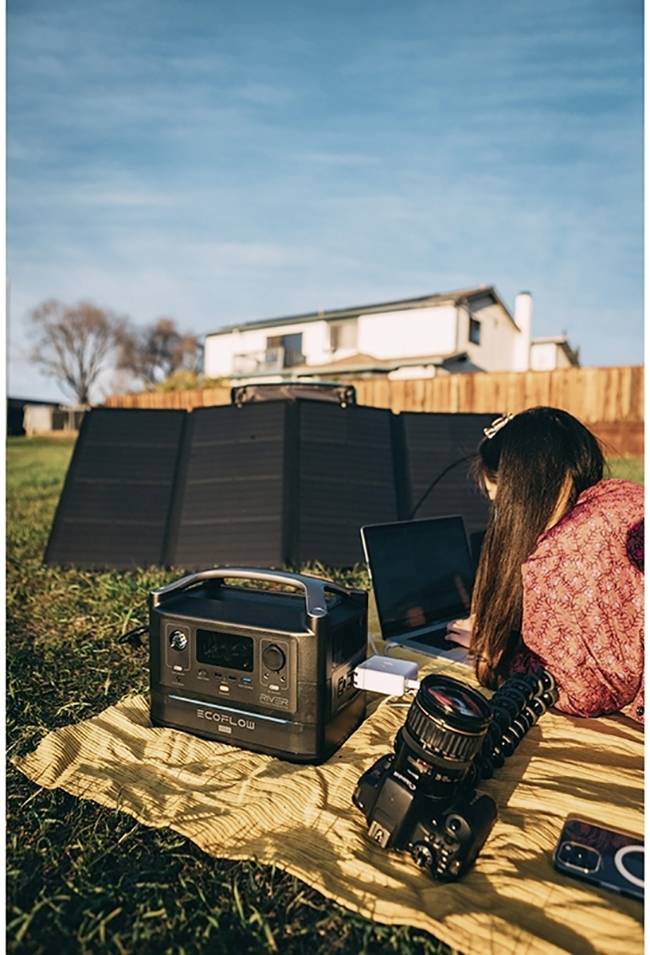 A person is sitting on a blanket outdoors, working on a laptop, with a portable power station and a camera nearby. Solar panels and a house are visible in the background.