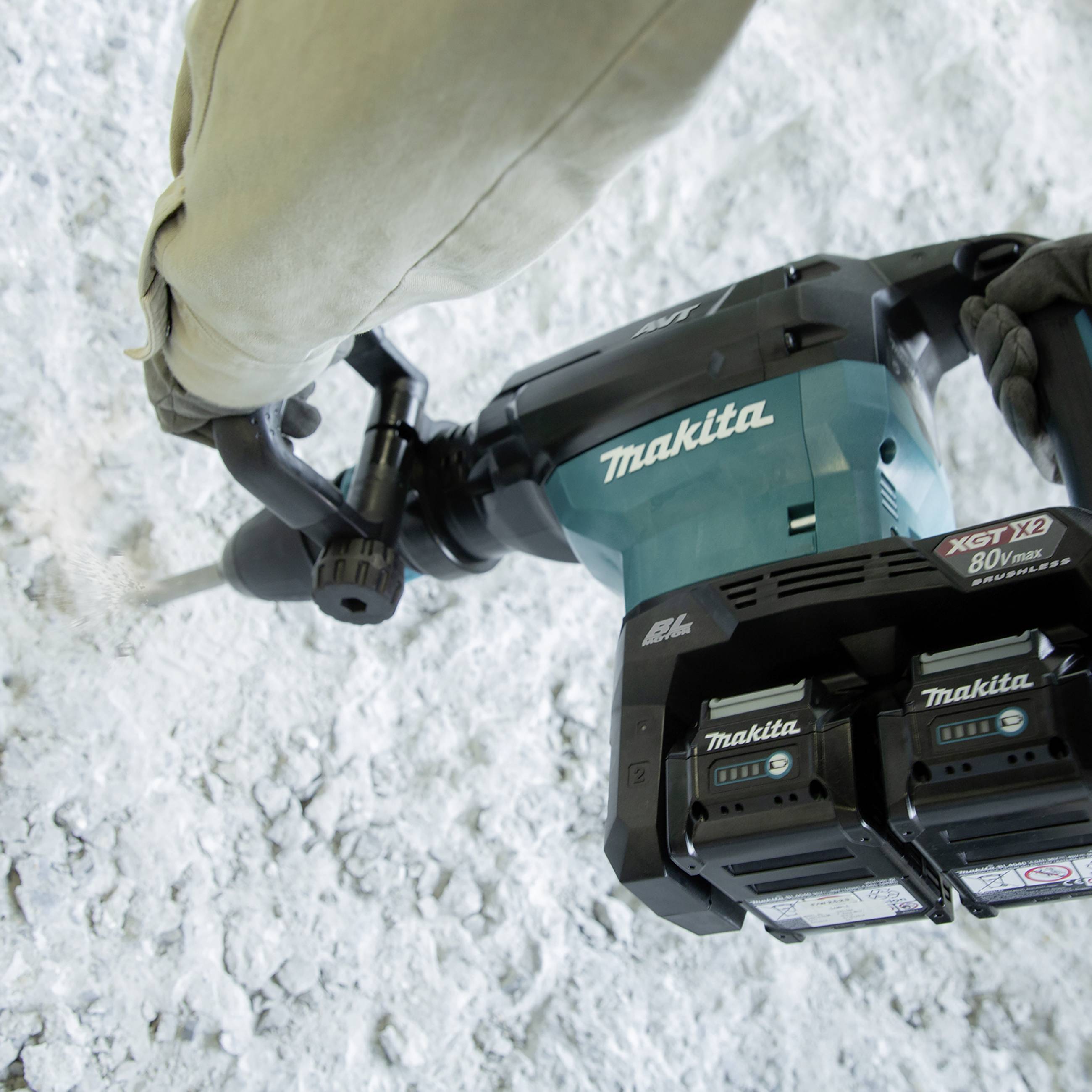 A person is using a Makita demolition hammer on a concrete surface. The device has two visible batteries.