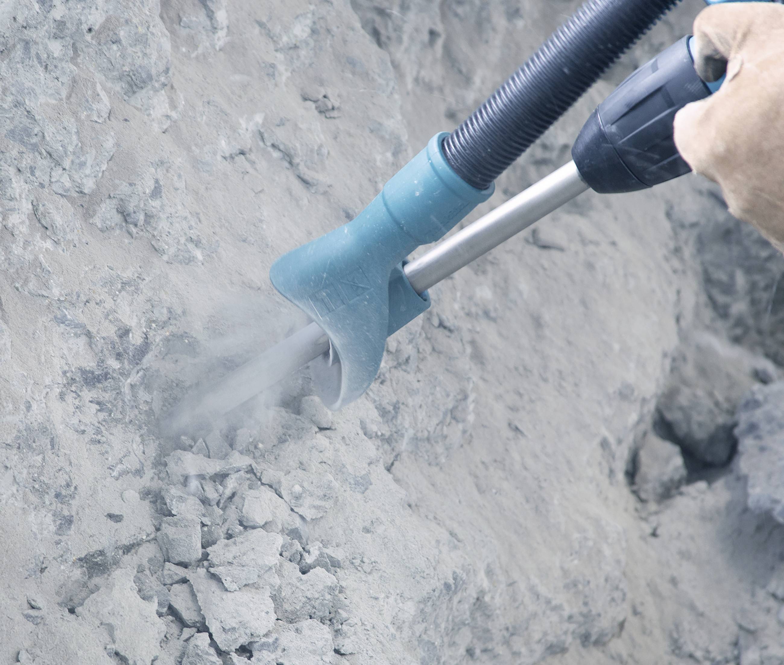 A hammer drill is working on a concrete wall. Dust is being extracted. The focus is on the tool and the work area.