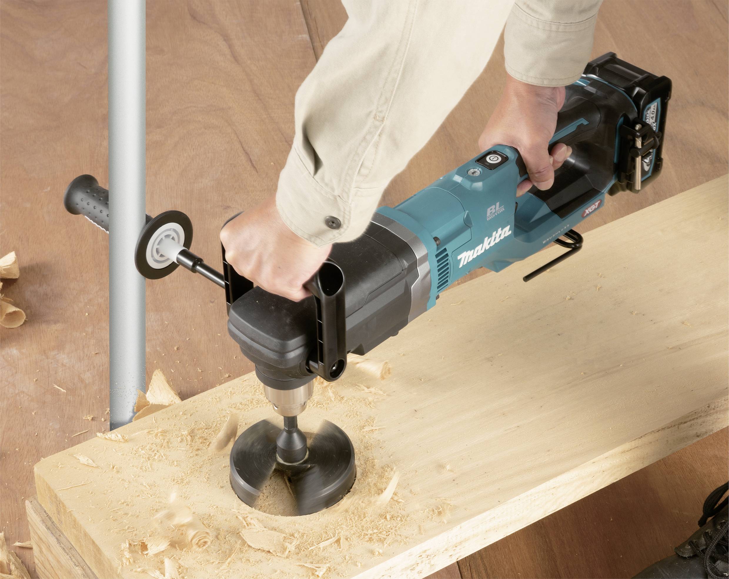 A person is using an electric hand drill to bore a large hole in a wooden board. Wood shavings are visible.