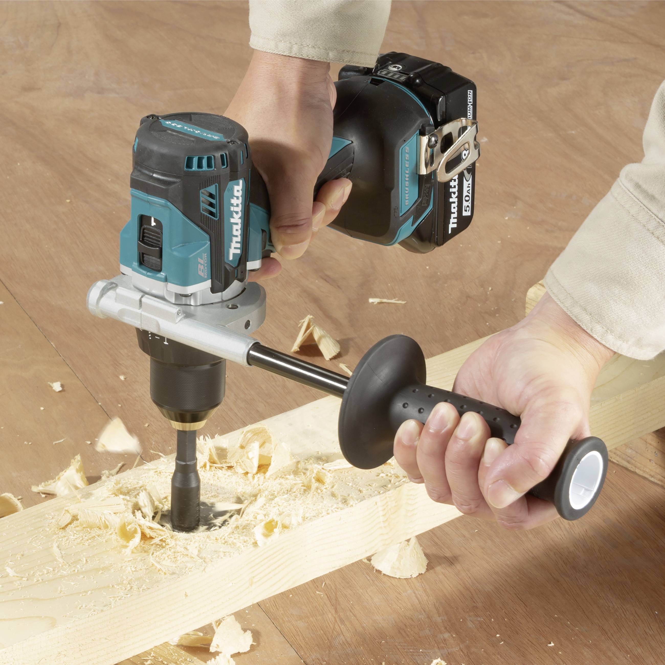 A person is using a blue cordless drill with a drilling attachment to drill a hole in a wooden board. Wood shavings are visible.