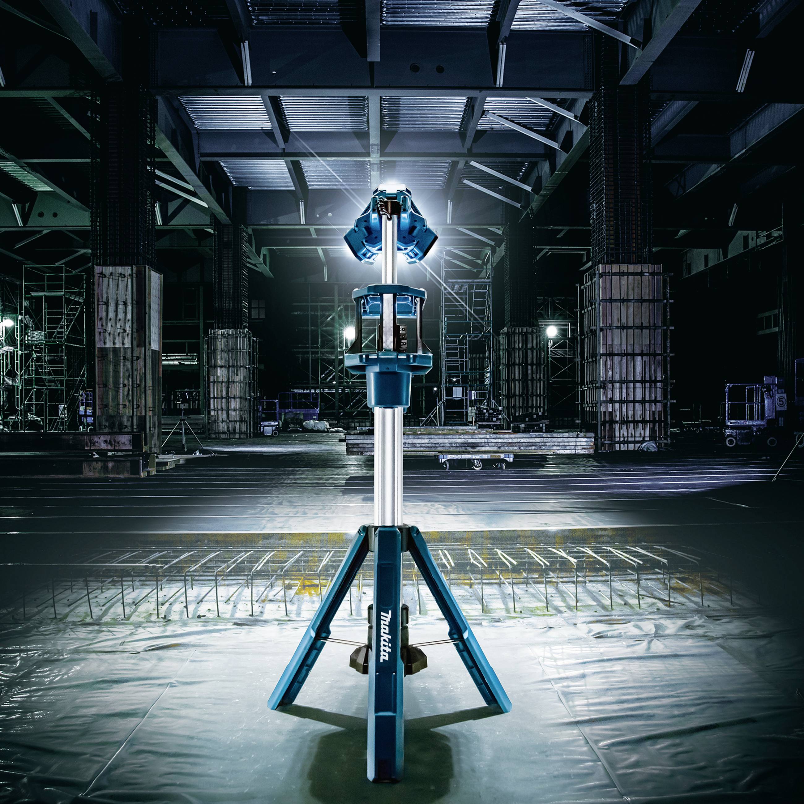 A work light stands on a tripod in a large, industrial hall. The floodlight illuminates the dark space.