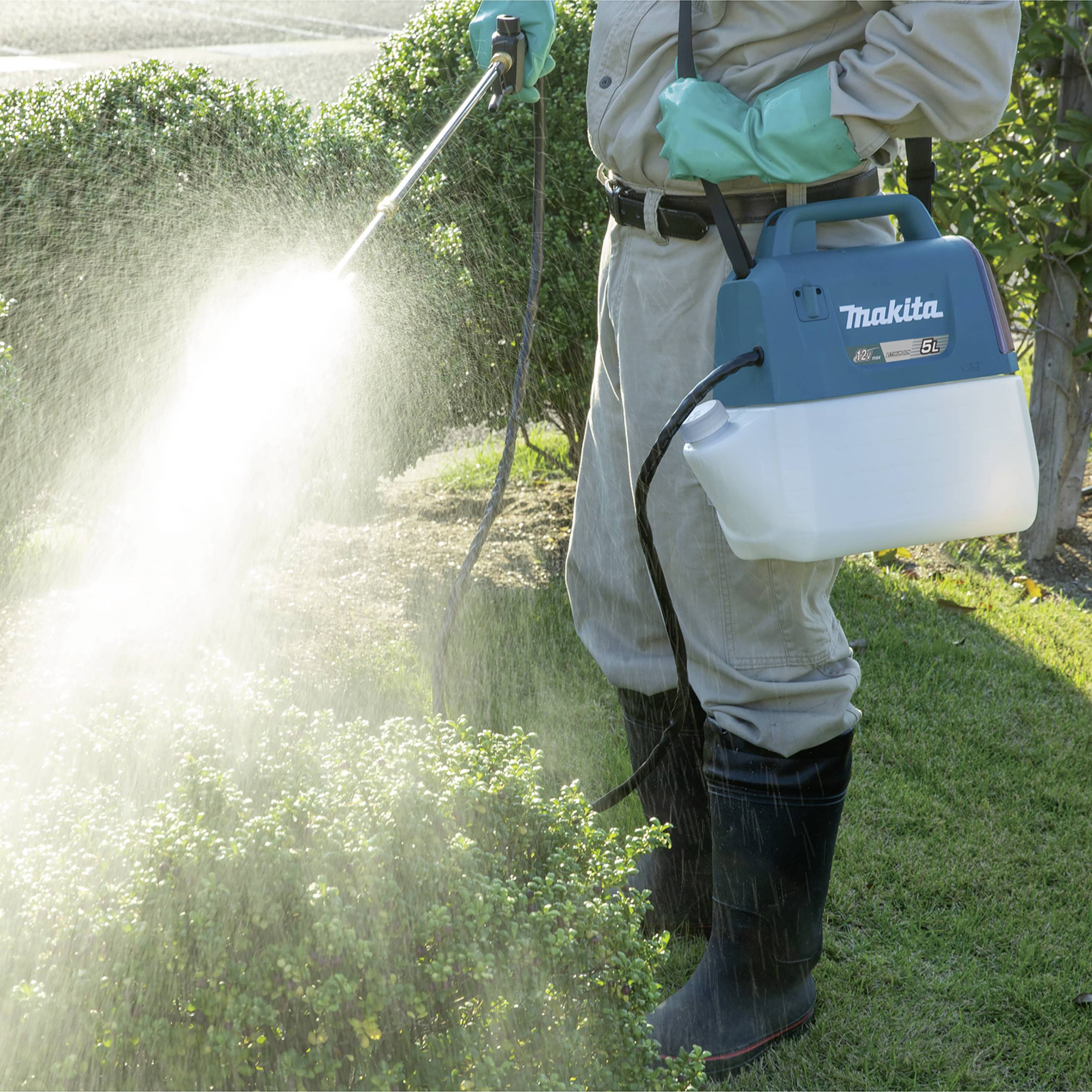 A person is spraying pesticide on bushes in a garden. They are wearing protective clothing with gloves and boots.