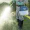 A person is spraying pesticide on bushes in a garden. They are wearing protective clothing with gloves and boots.