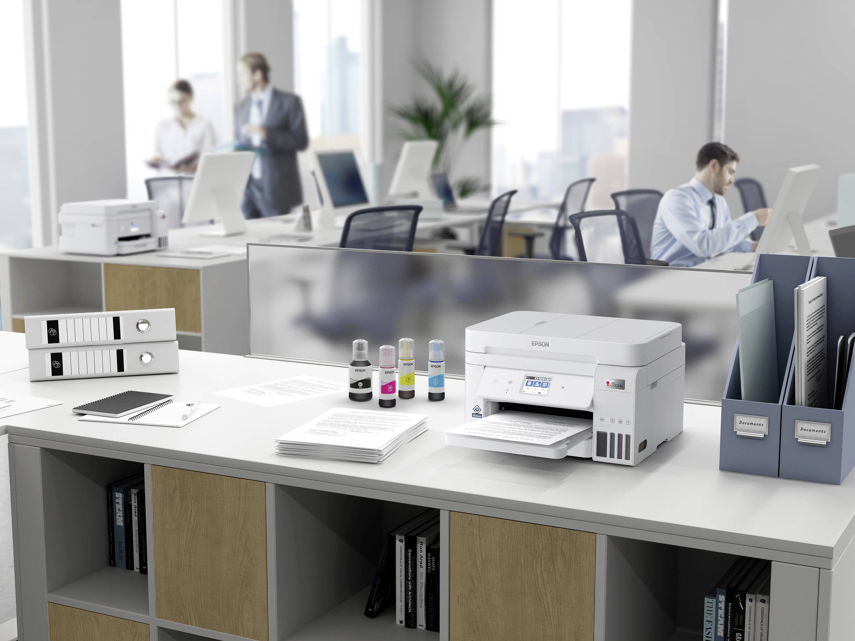 A modern office with computers on desks. In the foreground stands a printer, surrounded by ink and documents, with employees in the background.