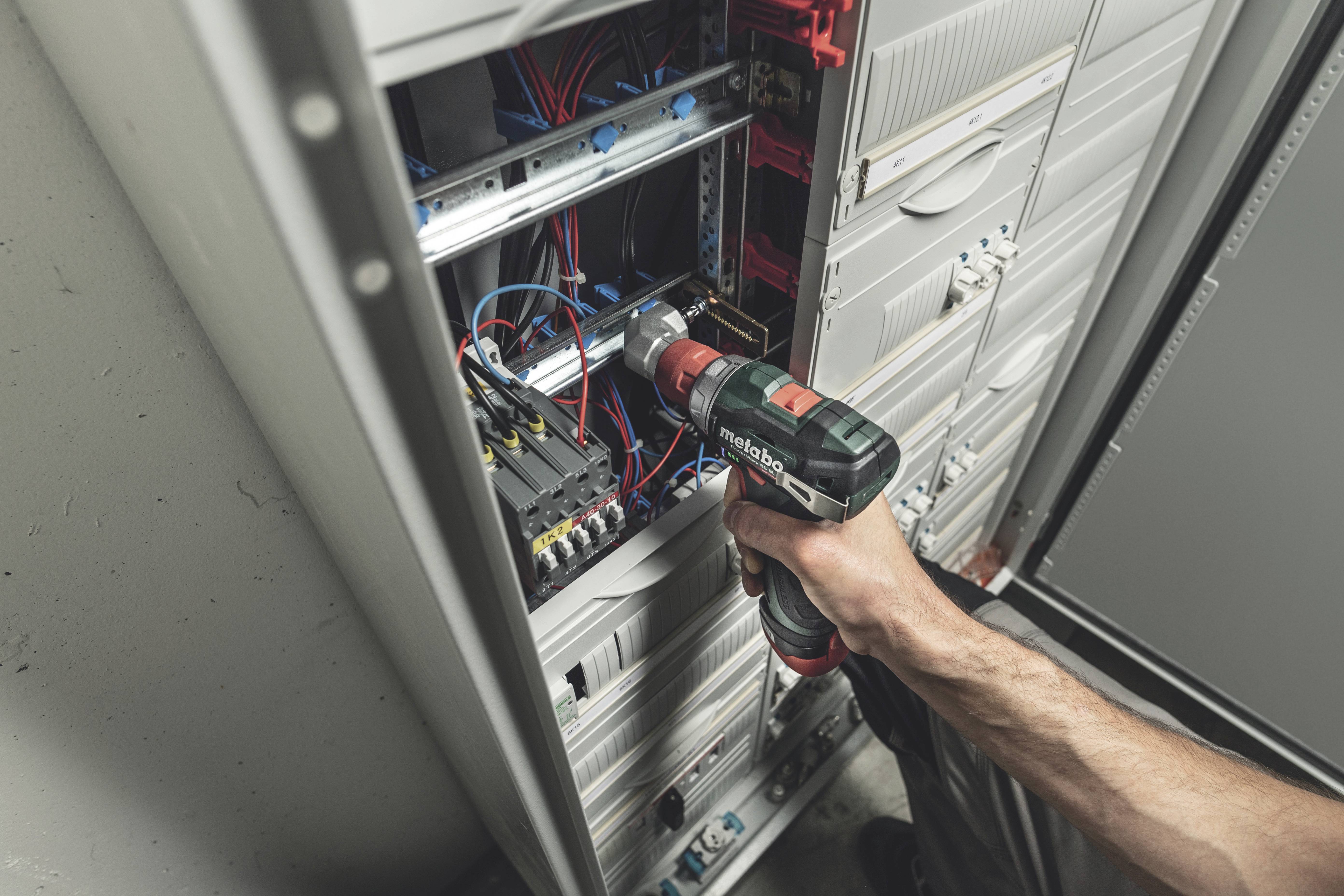 A person is using a cordless drill to work on an open electrical cabinet, with numerous colourful cables visible inside.