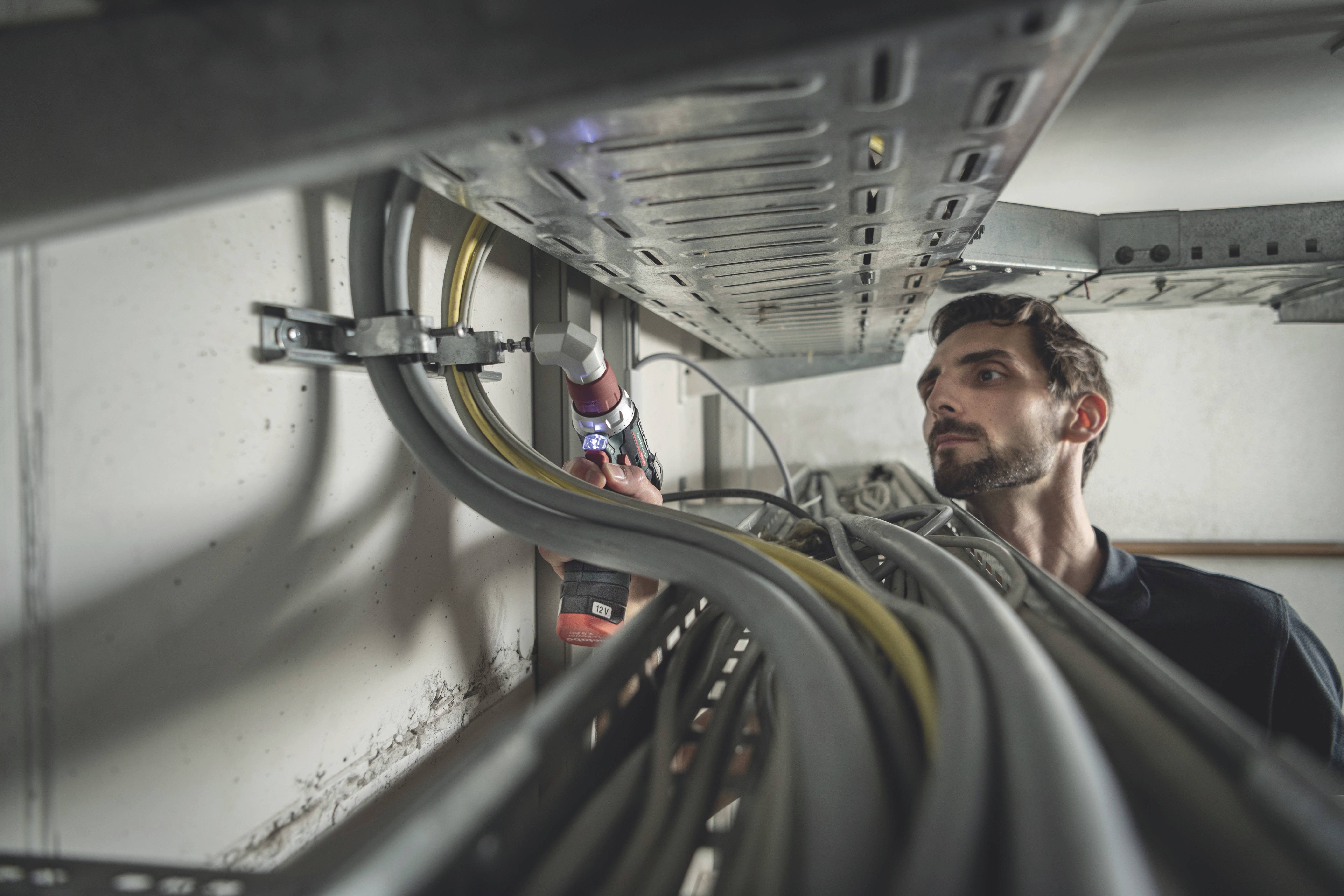 A man is working on a cable trunking system in a technical room, using an electric tool. Cables run along the wall.