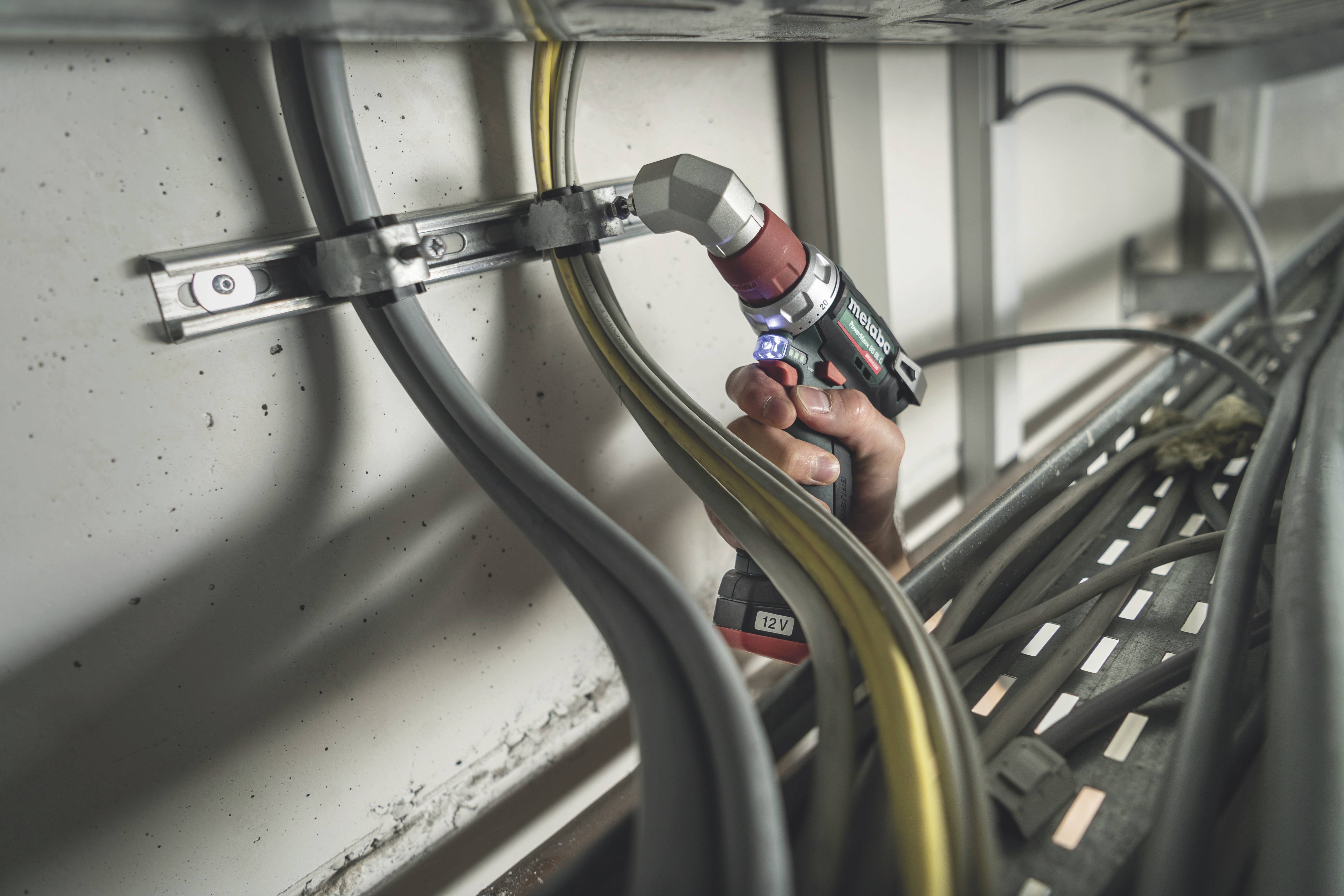 A hand holds a drill and tightens cables within a metal frame. Various cables run parallel along the wall.