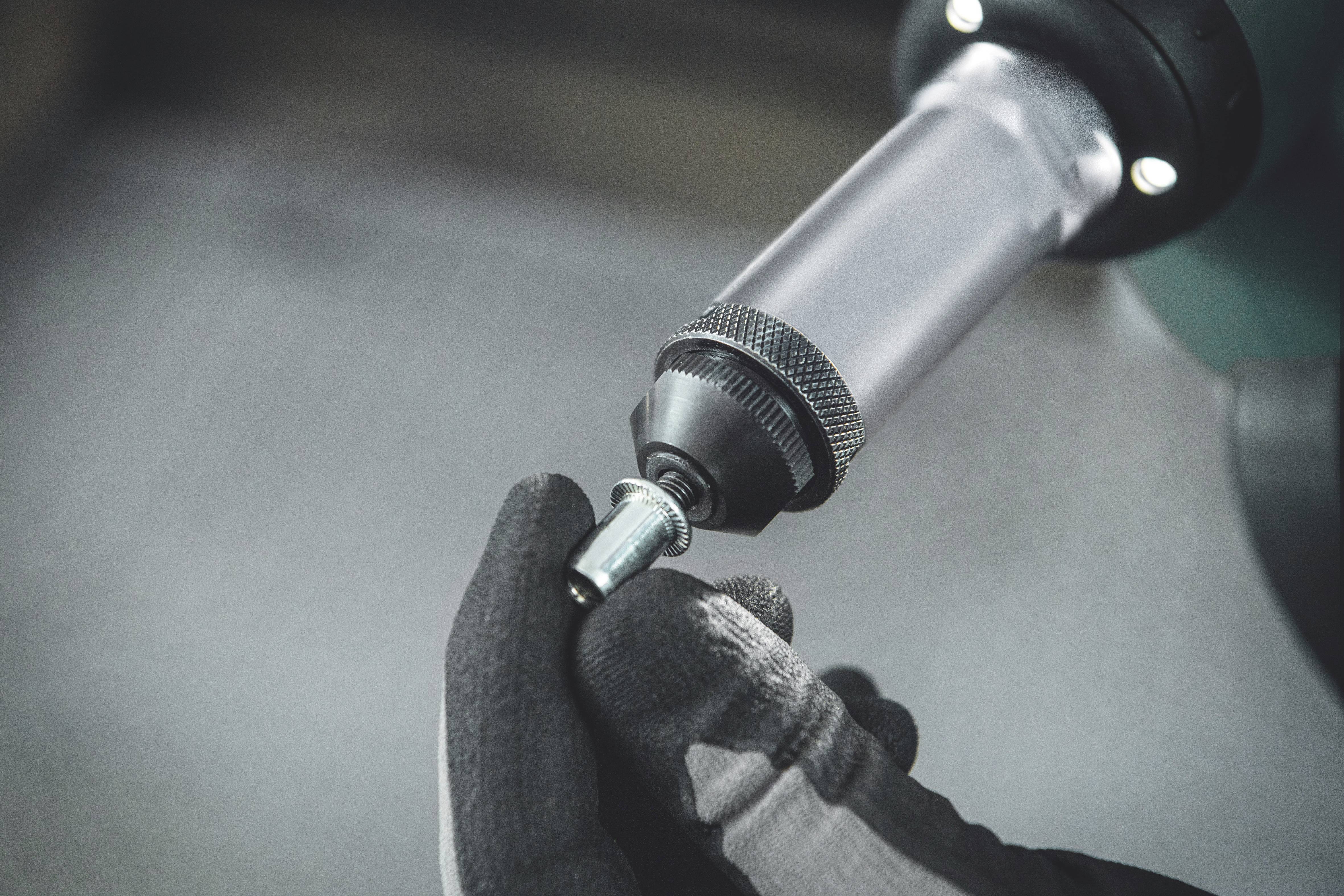 A gloved hand holds a small screw while an electric tool tightens the screw.