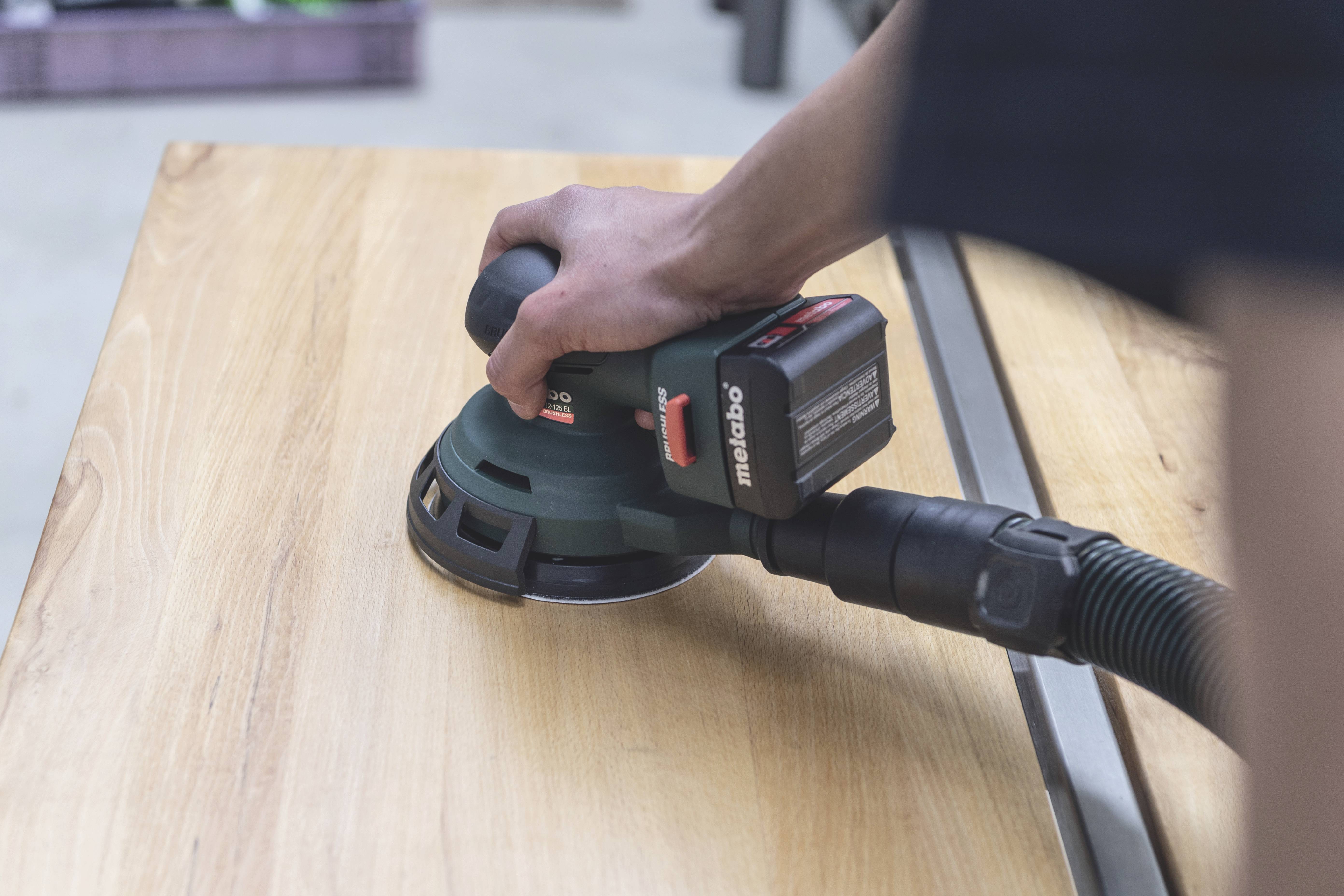 A person is sanding a wooden board with an electric sanding tool. Dust is being extracted through an attached hose.