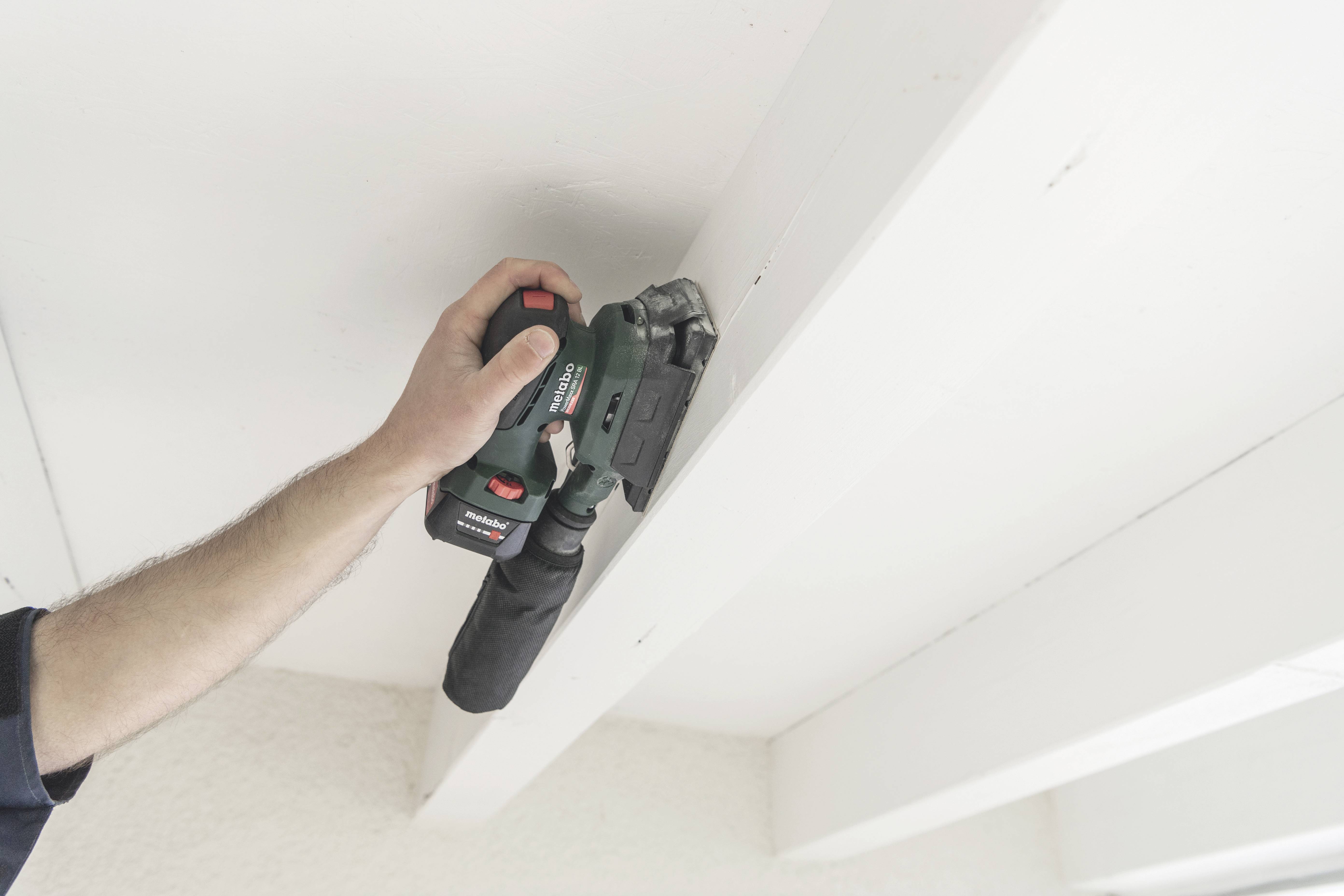 A hand holds an electric sanding tool, working on a wooden beam to smooth the surface.