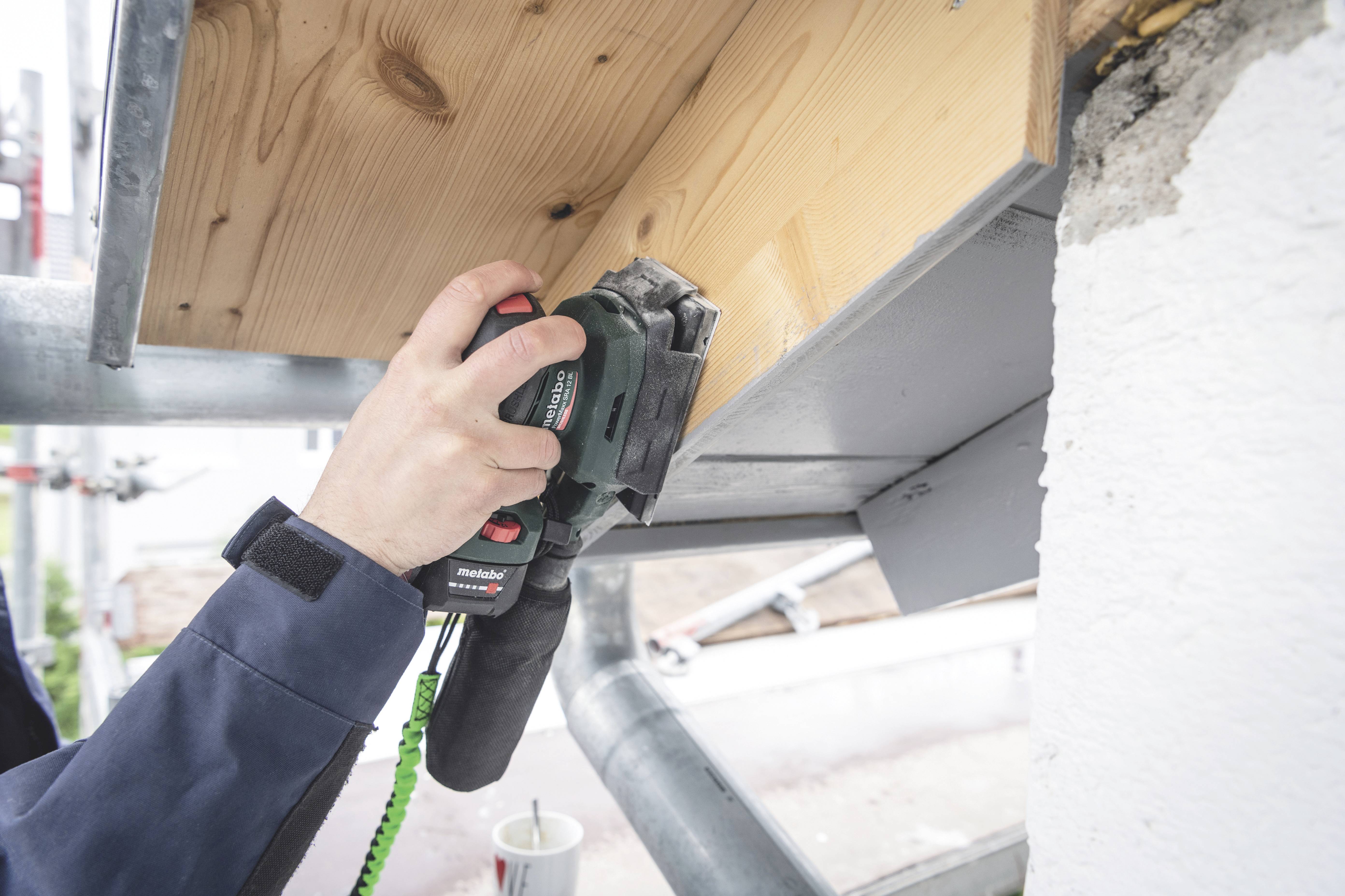 A person is sanding wood with an electric sanding tool on an eaves overhang. The scene depicts craftsmanship outdoors.